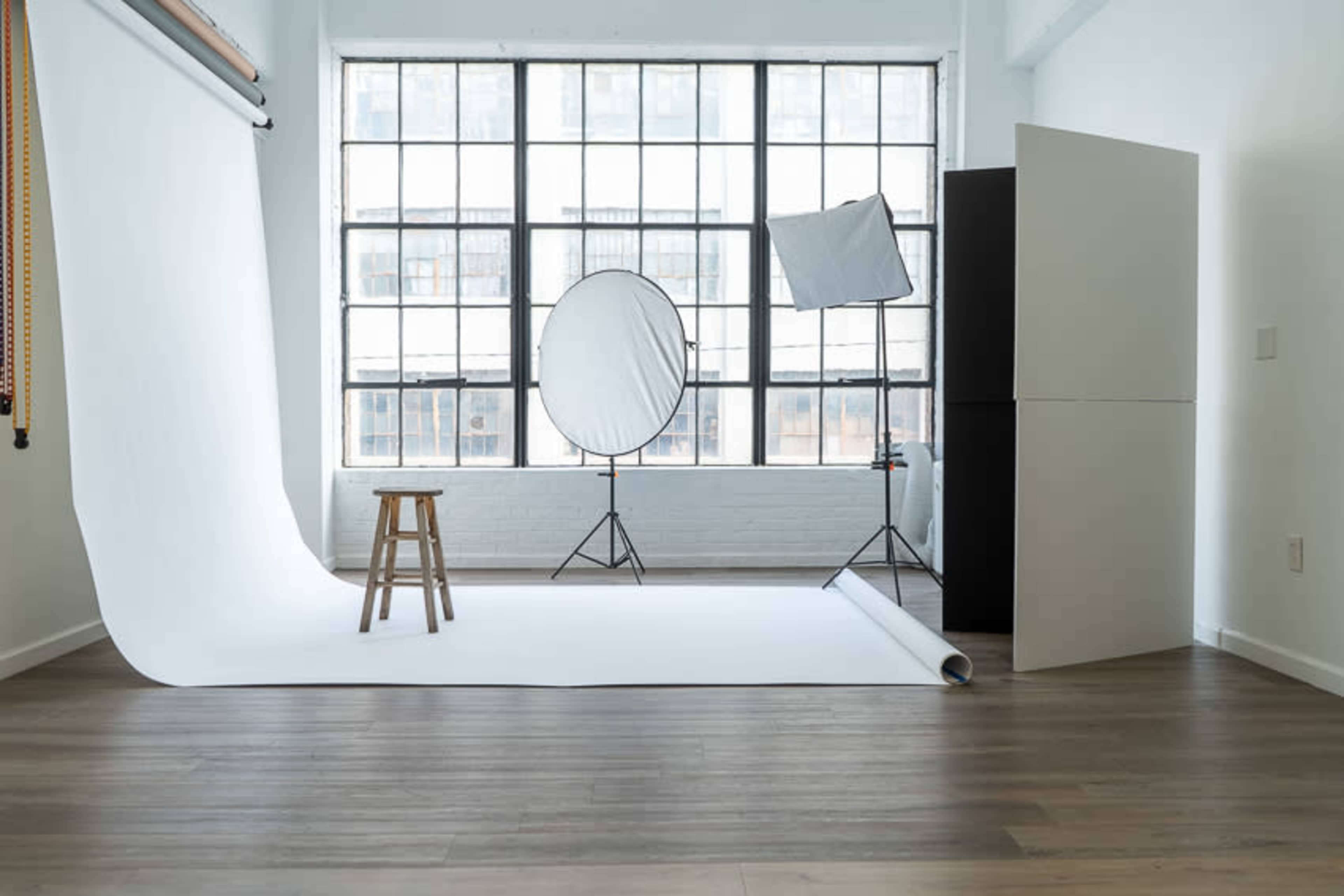 A photography studio features a white backdrop, a stool, and lighting equipment set up near a large window.