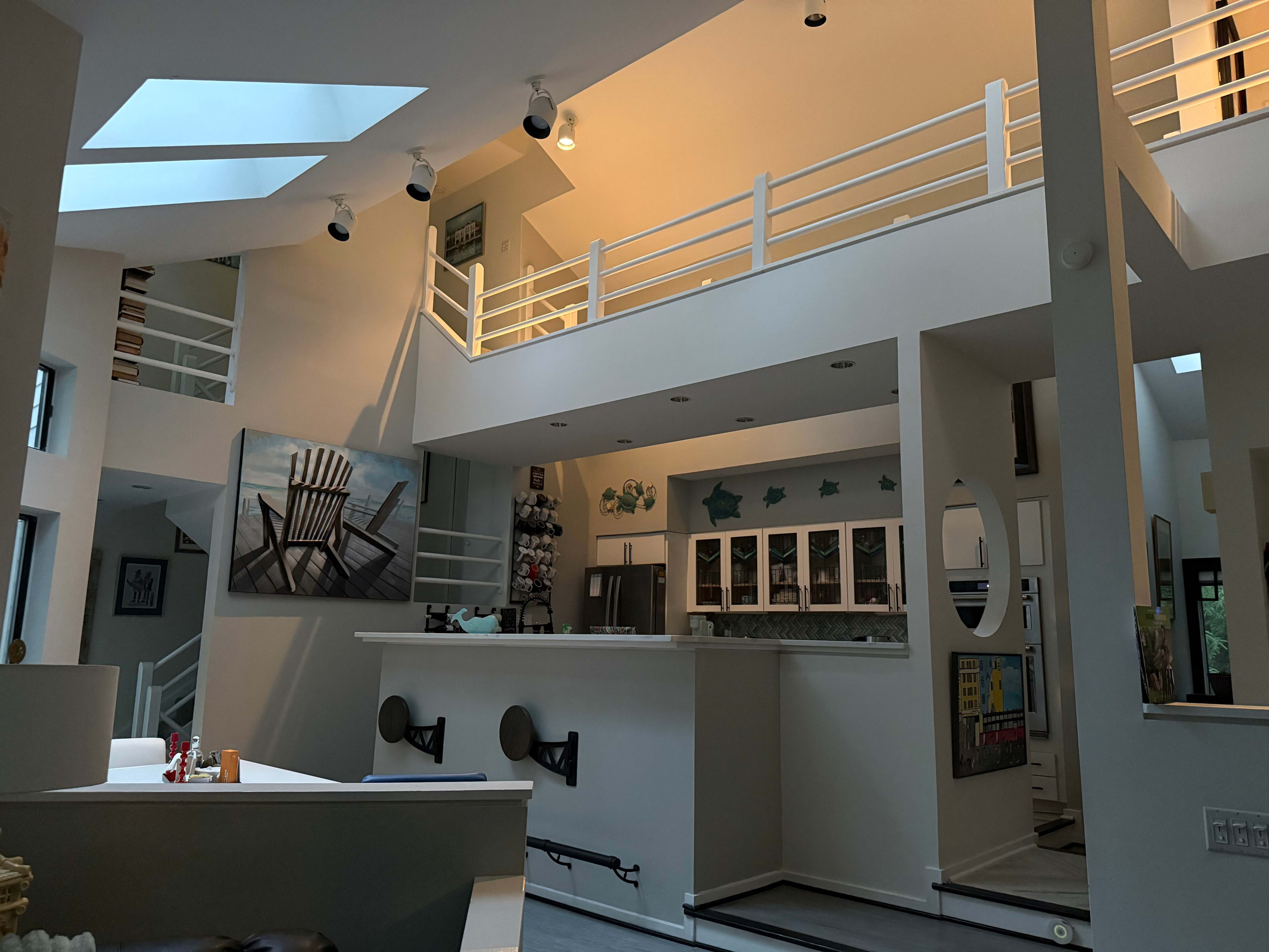 The image shows a modern, open-concept living space with high ceilings, large windows, and a balcony overlooking the lower level.