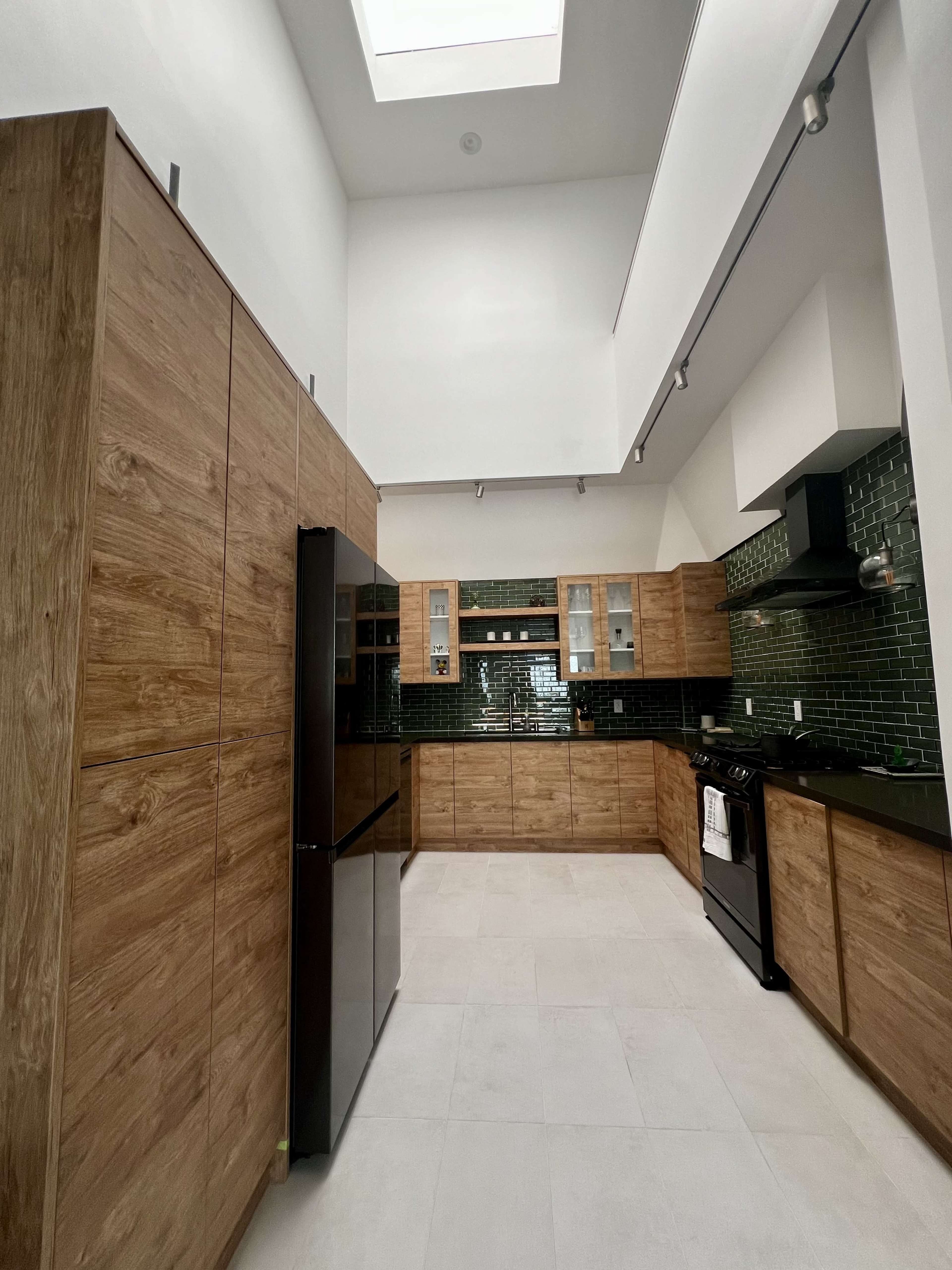 A modern kitchen features wood cabinetry, black appliances, and green tiled backsplash under a high ceiling with a skylight.