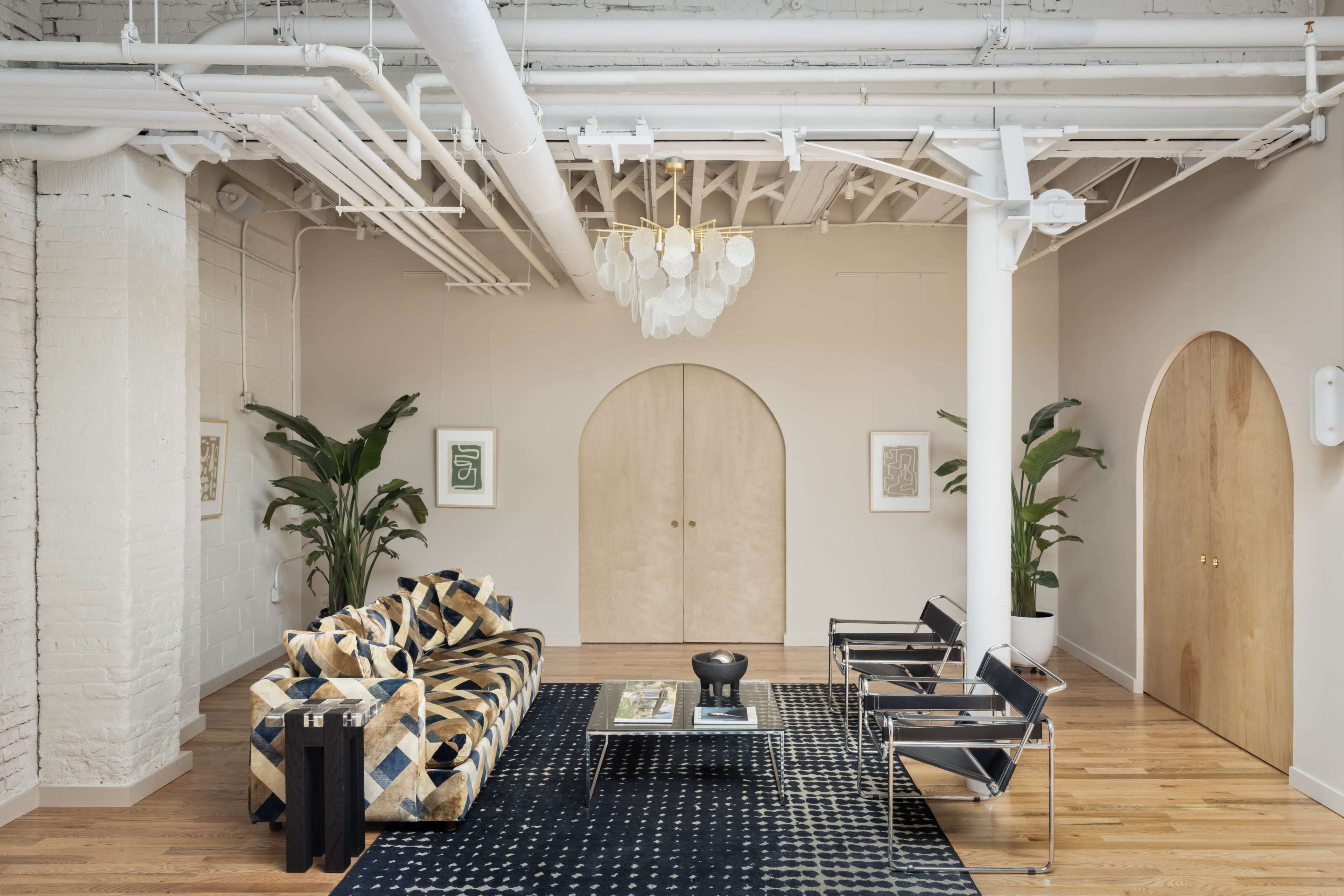 A modern living space featuring a patterned sofa, a black coffee table, and two contemporary chairs, with plants and arched doorways in the background.
