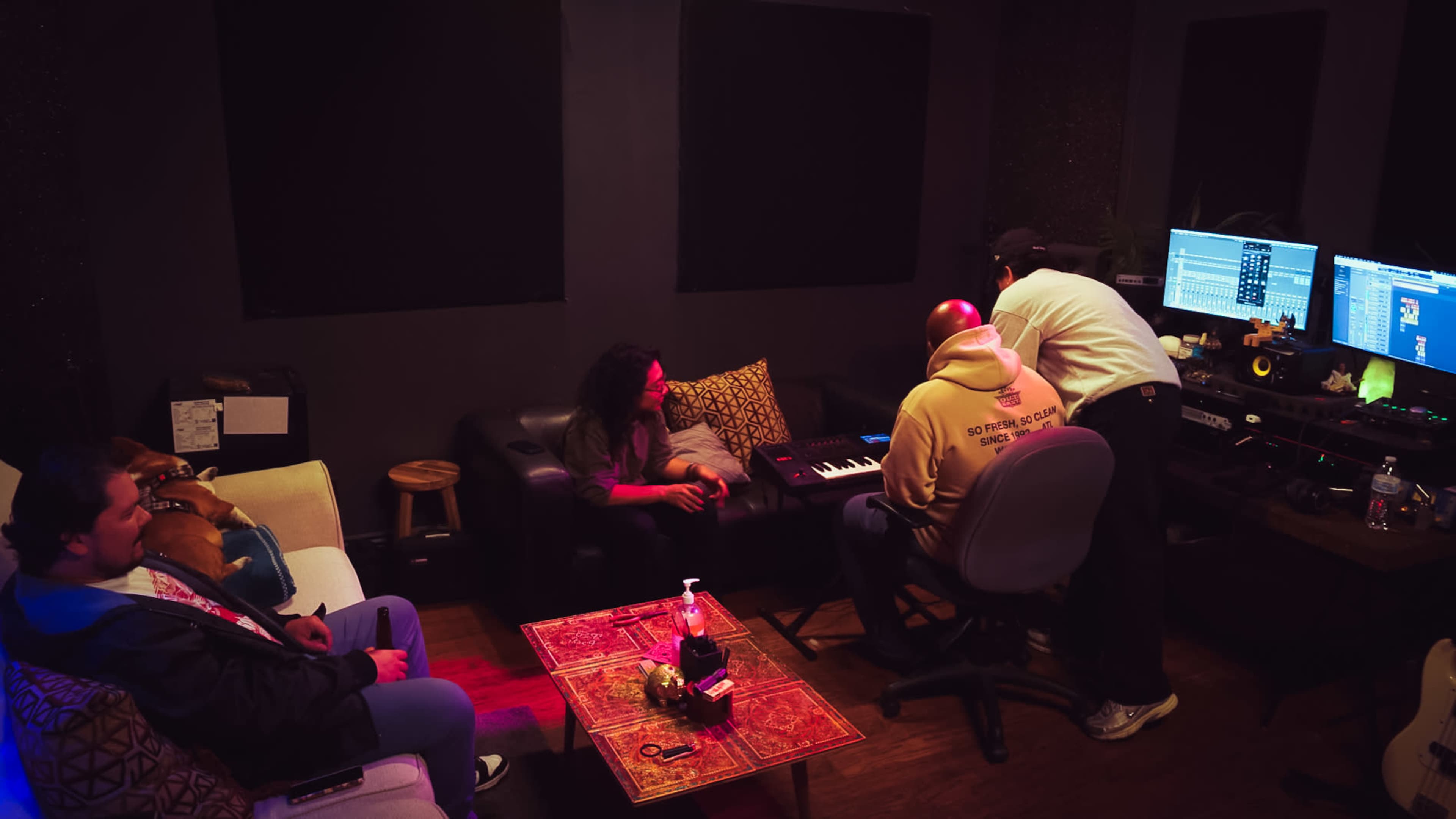 A group of four people is working in a music studio, with one person playing a keyboard while others observe and discuss the process.