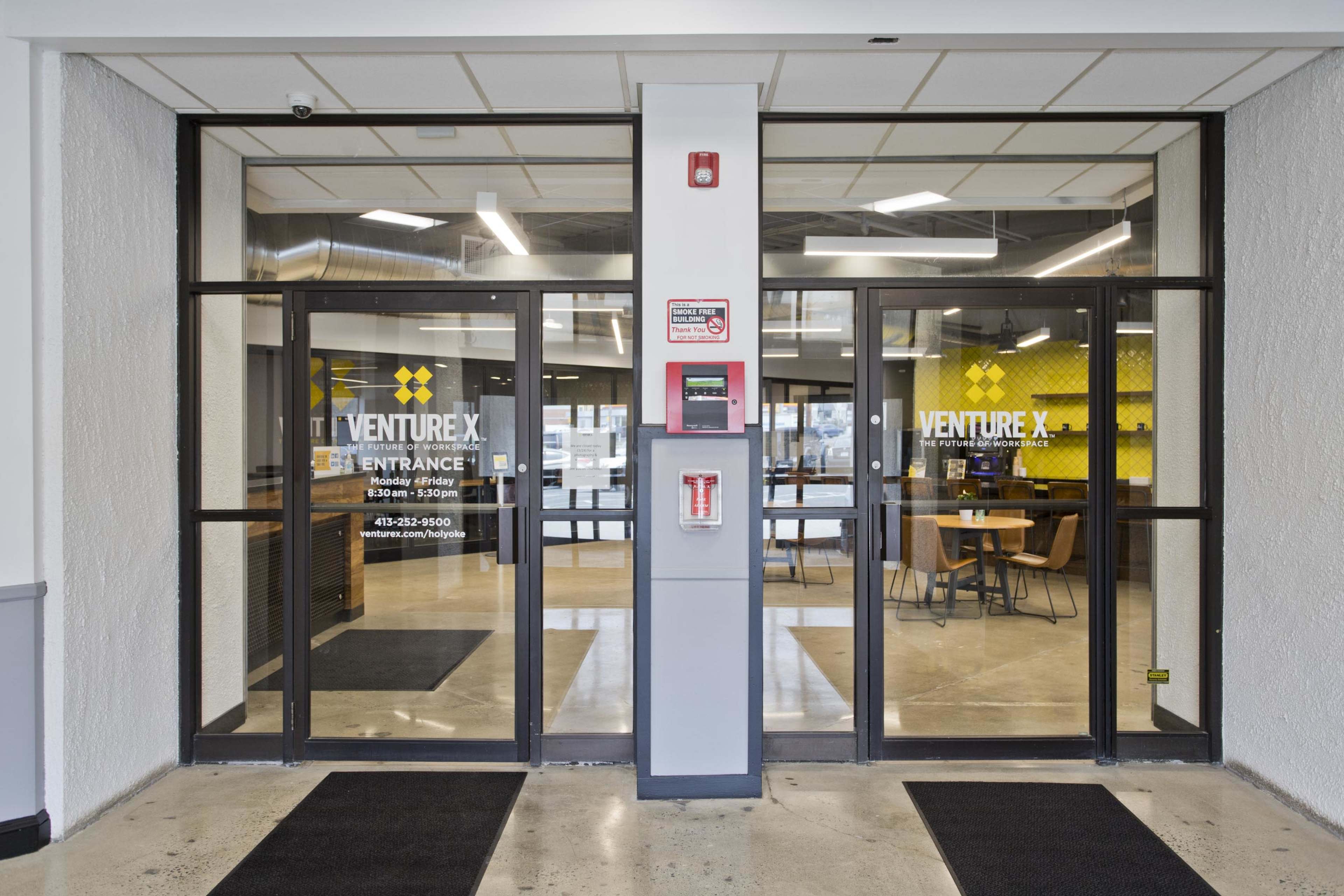 The entrance of a shared workspace called Venture X, featuring glass double doors leading into a modern office area with yellow accents and tables.