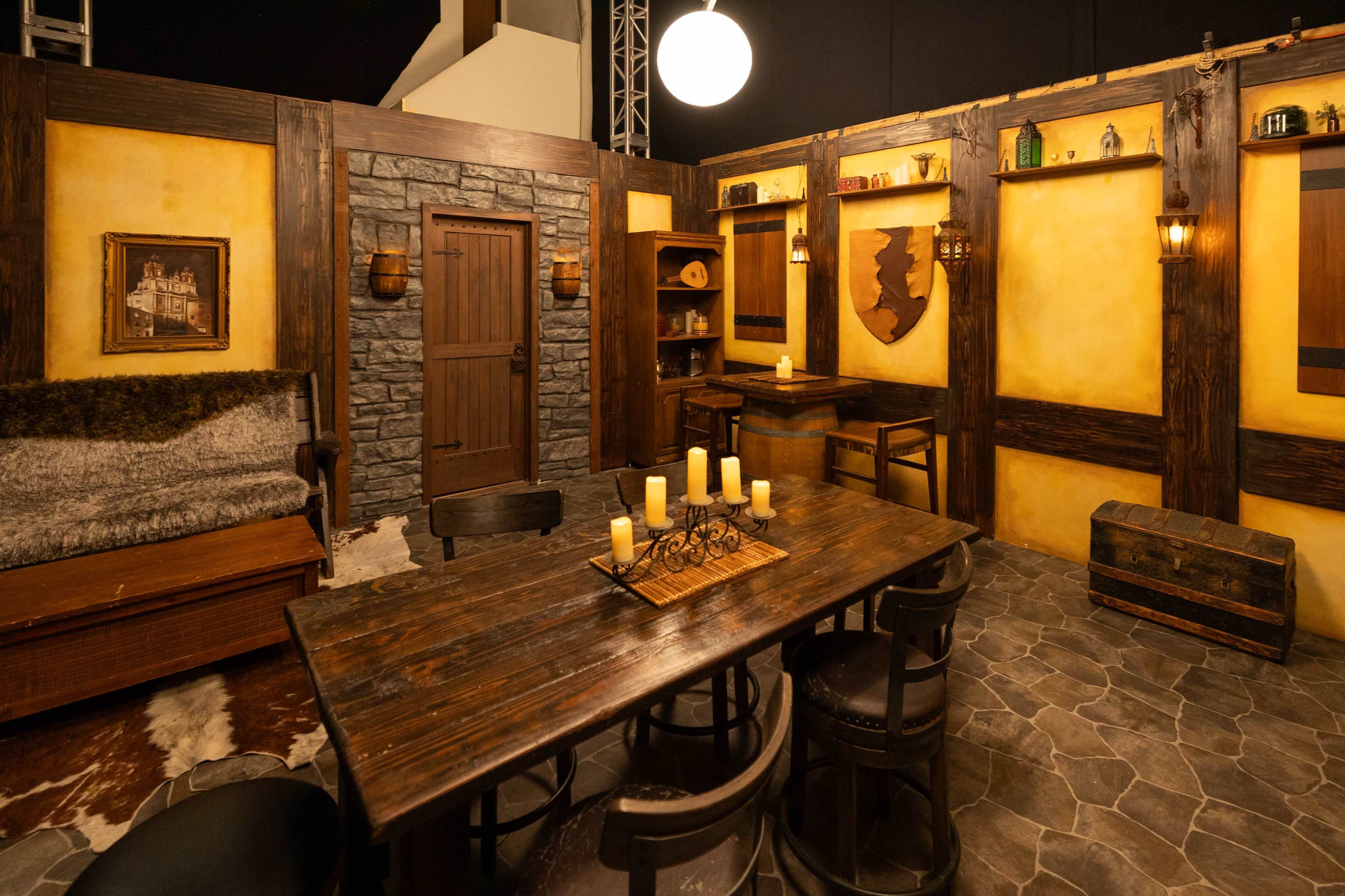 The image features a rustic-themed room with wooden beams, a stone wall, a wooden table surrounded by chairs, and flickering candles on the table.