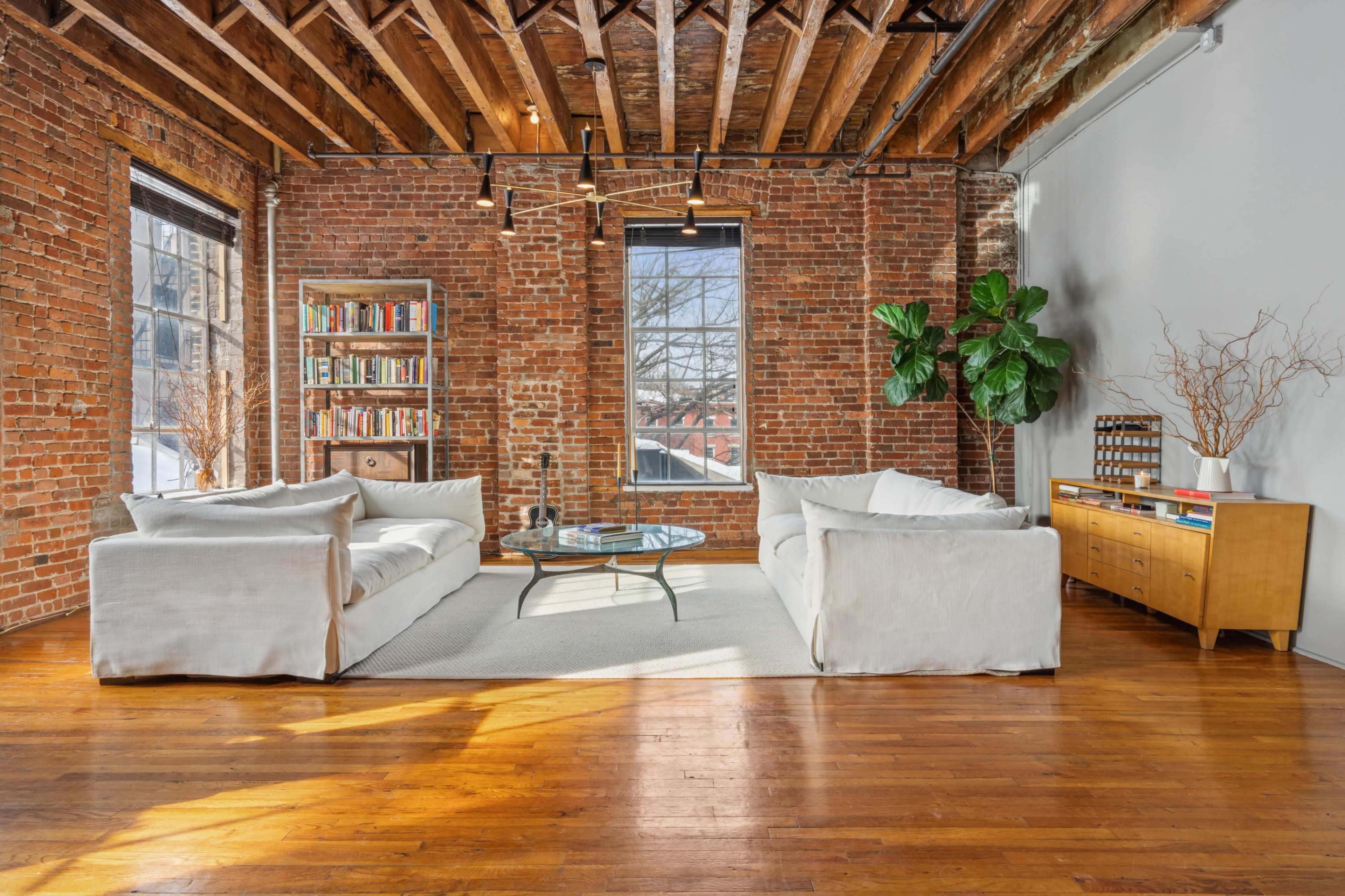 A bright living room features exposed brick walls, wooden beams, two white sofas, a coffee table, a bookshelf, and a sideboard with decorative items.