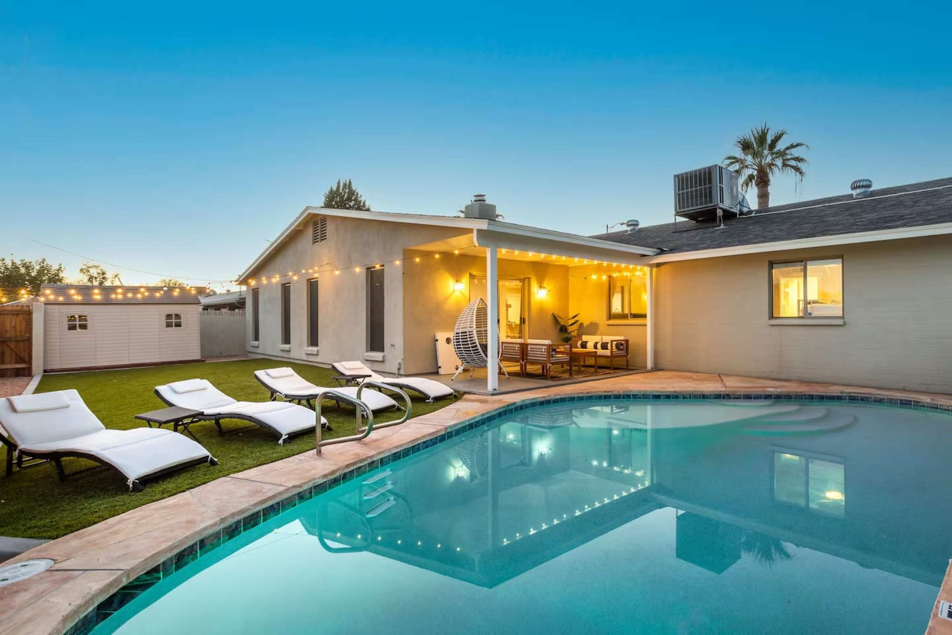 The image shows a backyard with a swimming pool, lounge chairs, and a house illuminated by string lights during twilight.