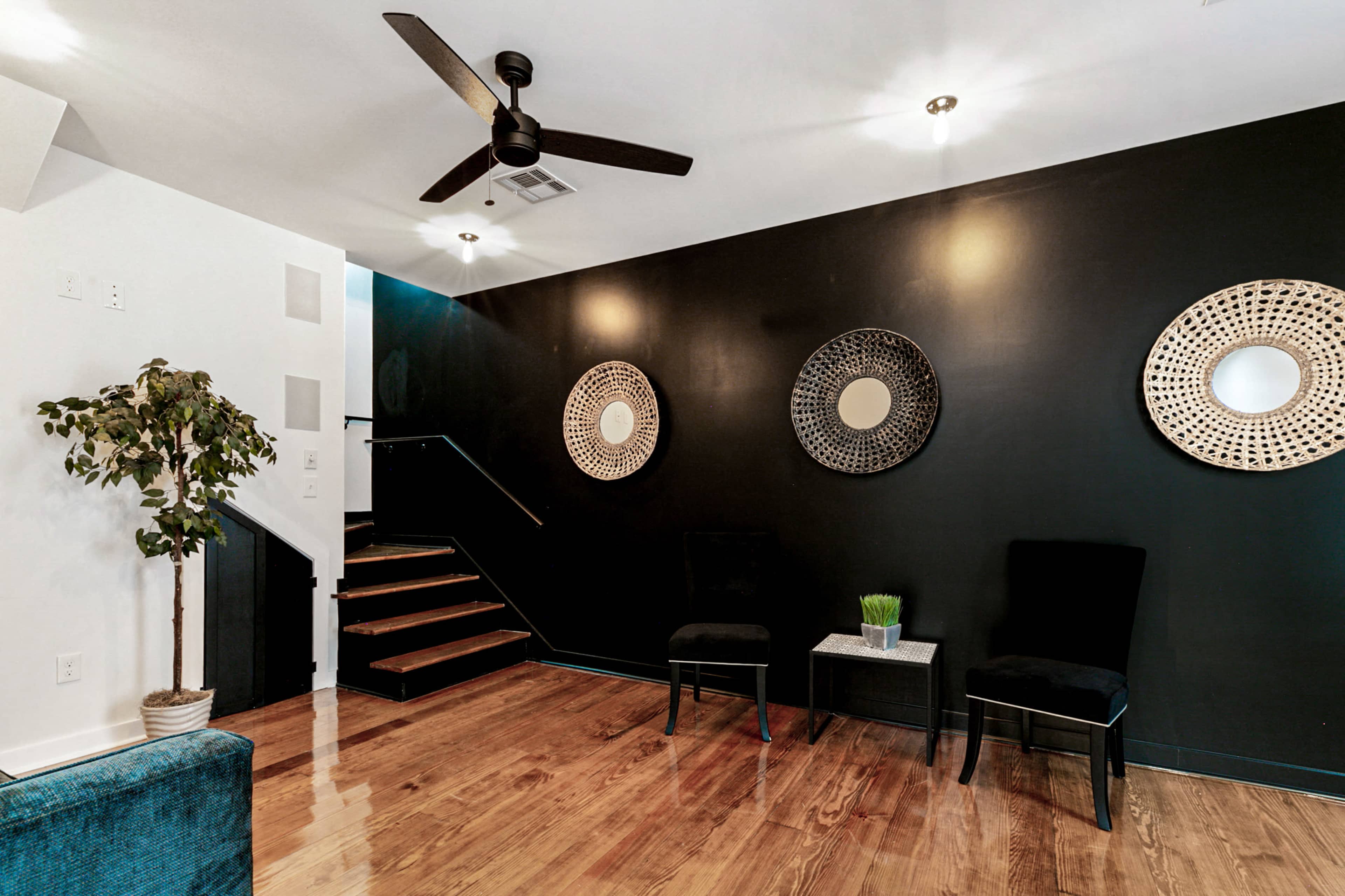 A modern interior space with dark walls, wooden floors, two black chairs, a small table with a plant, and a staircase leading to an upper level.