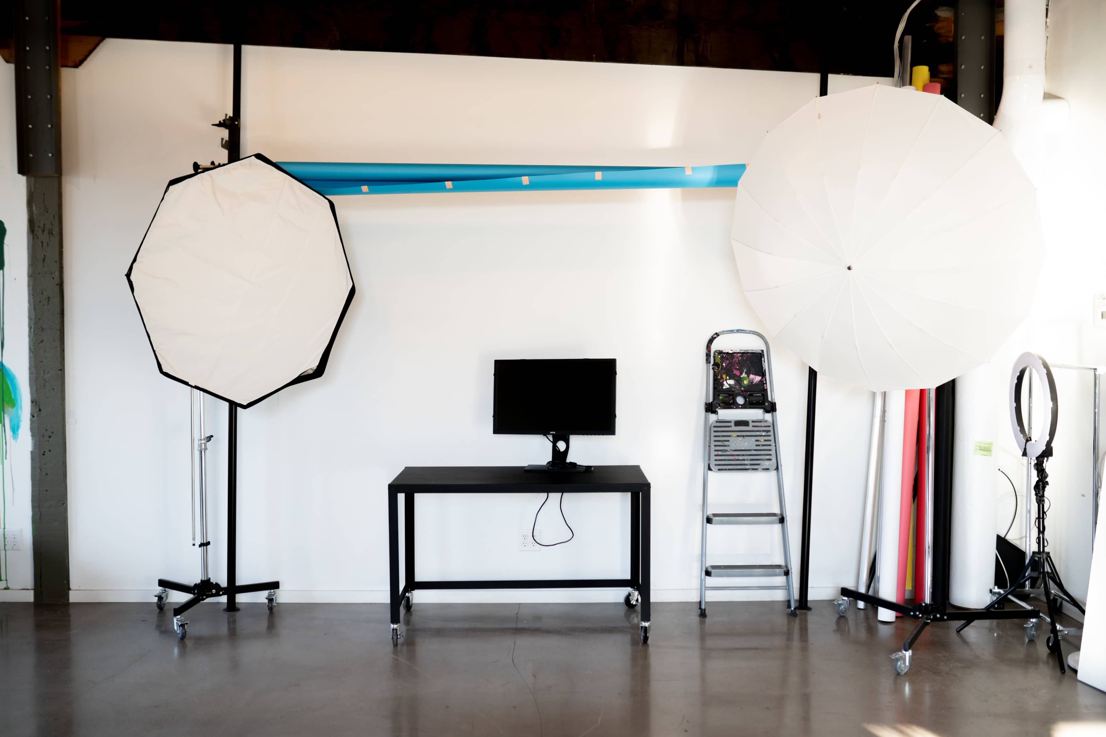 The image shows a studio setup featuring two large softbox lights, a black table with a monitor, a ladder, and several rolls of backdrop paper against a plain wall.