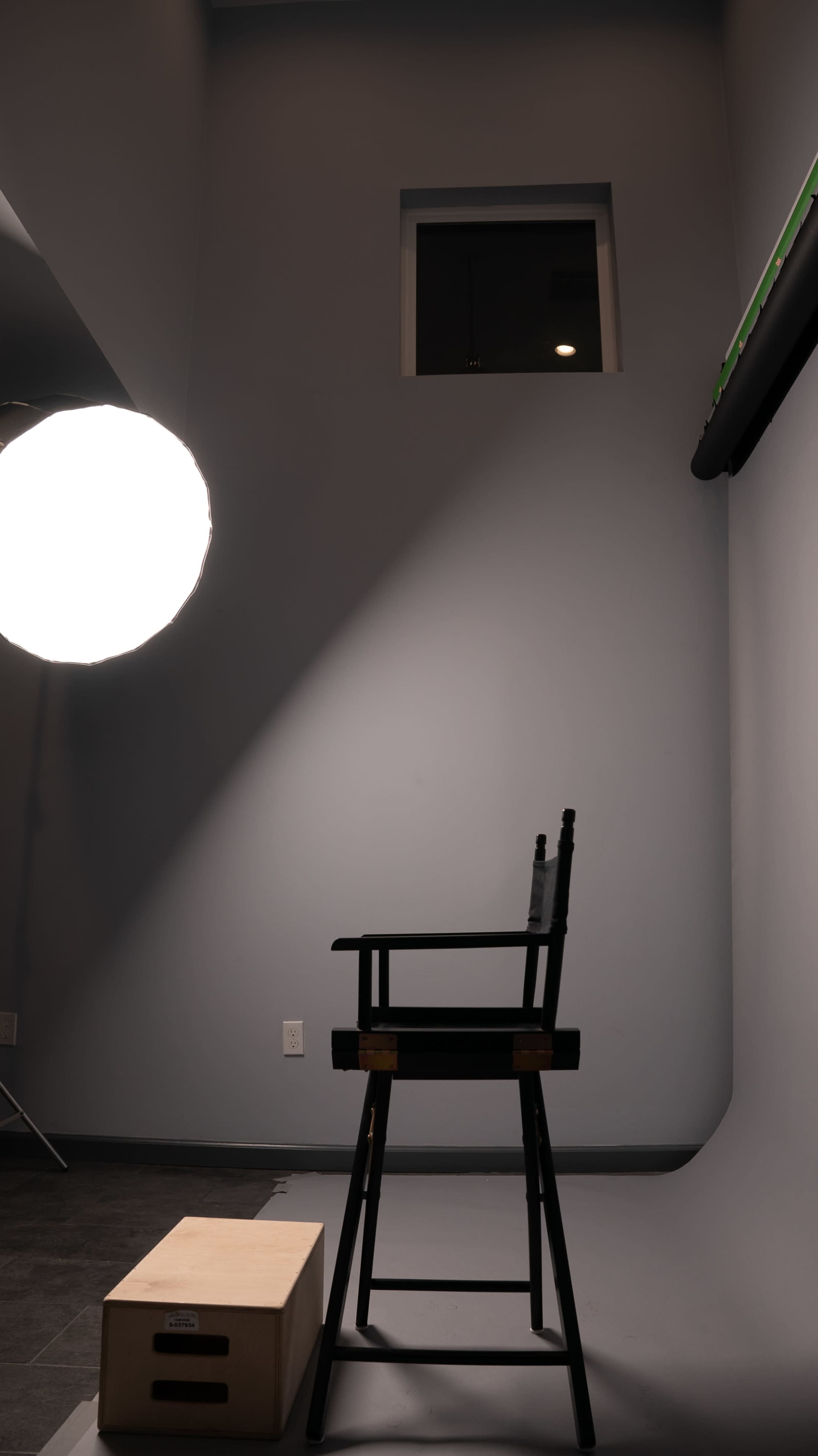 A simple wooden chair is positioned in front of a plain wall, illuminated by a large soft light source overhead.