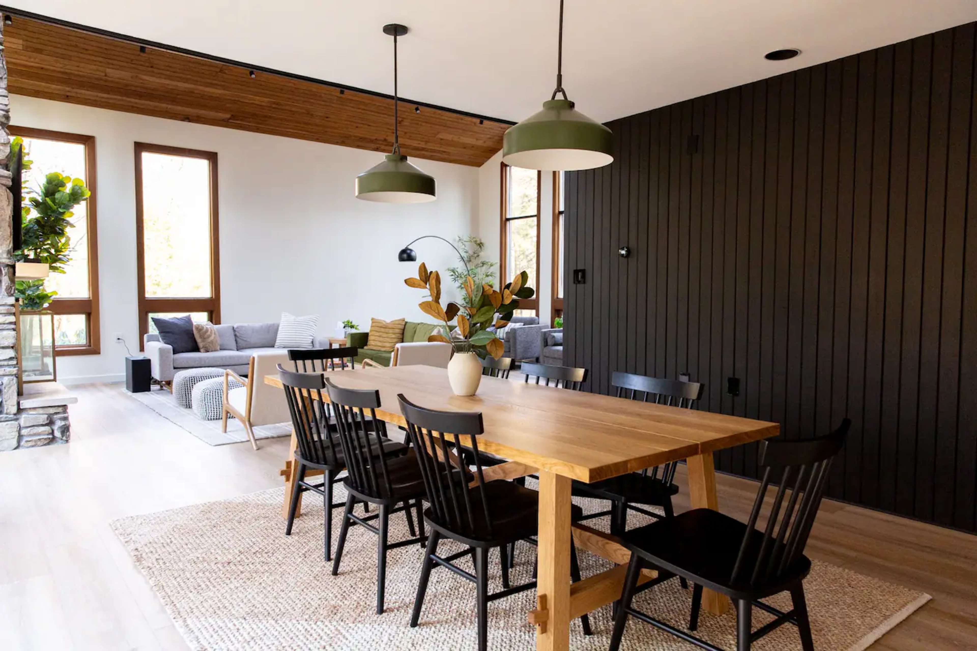 A modern dining area features a large wooden table surrounded by black chairs, with a cozy seating area and large windows in the background.