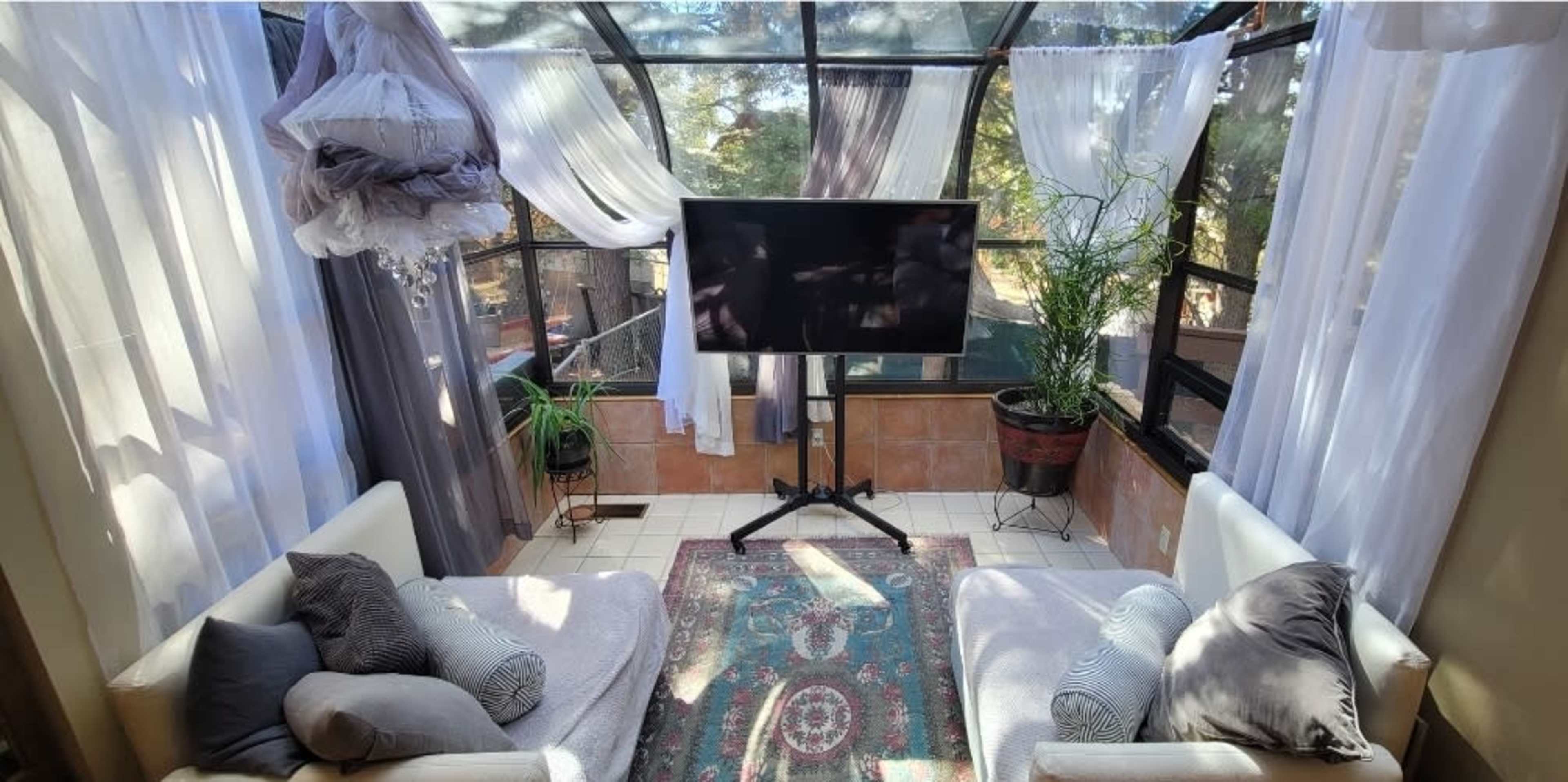 A sunlit sunroom features white curtains, two couches, a television on a stand, and potted plants.