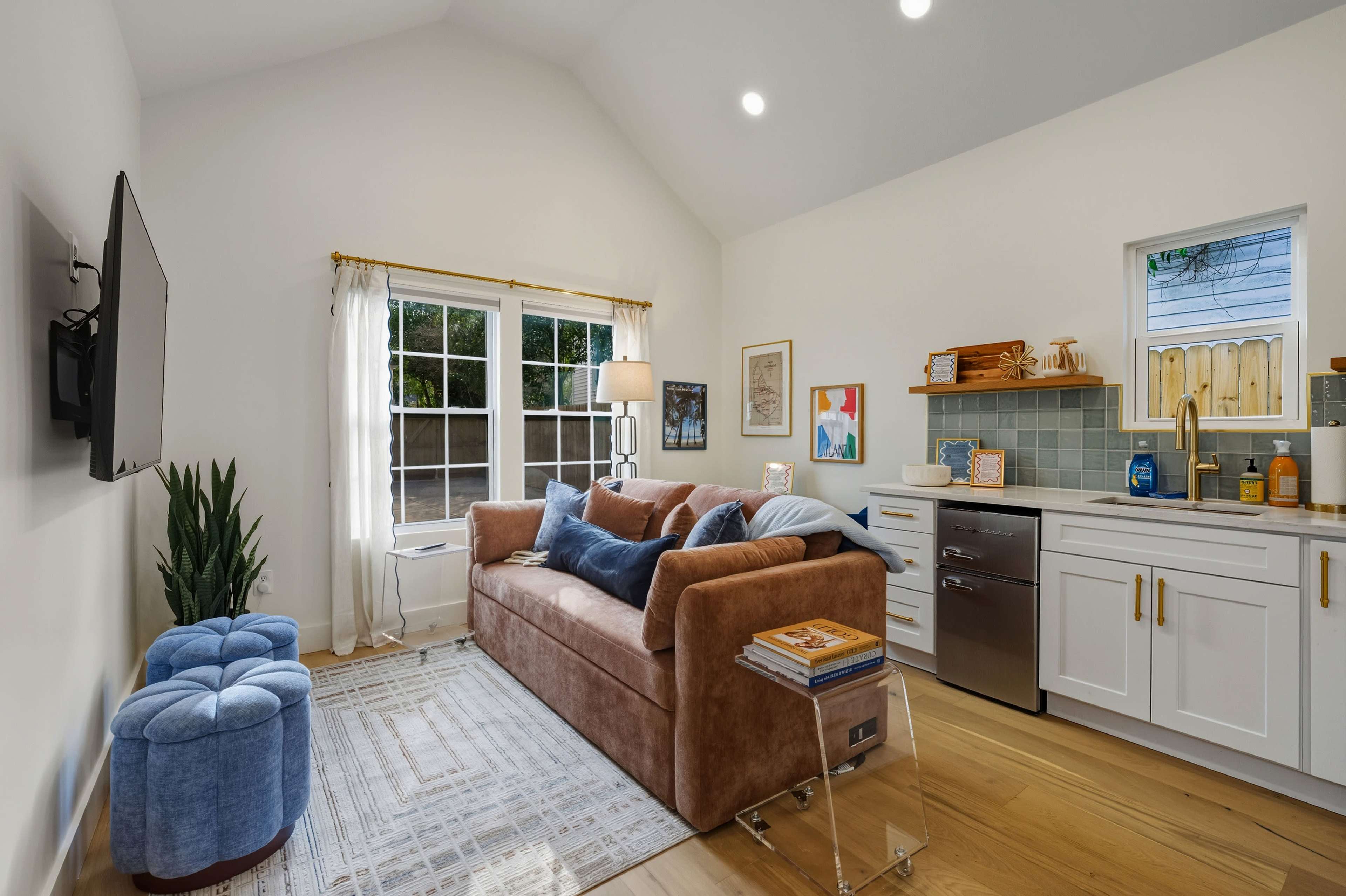 The image shows a cozy living space with a brown couch, a small blue ottoman, and a kitchenette in a well-lit room featuring large windows.