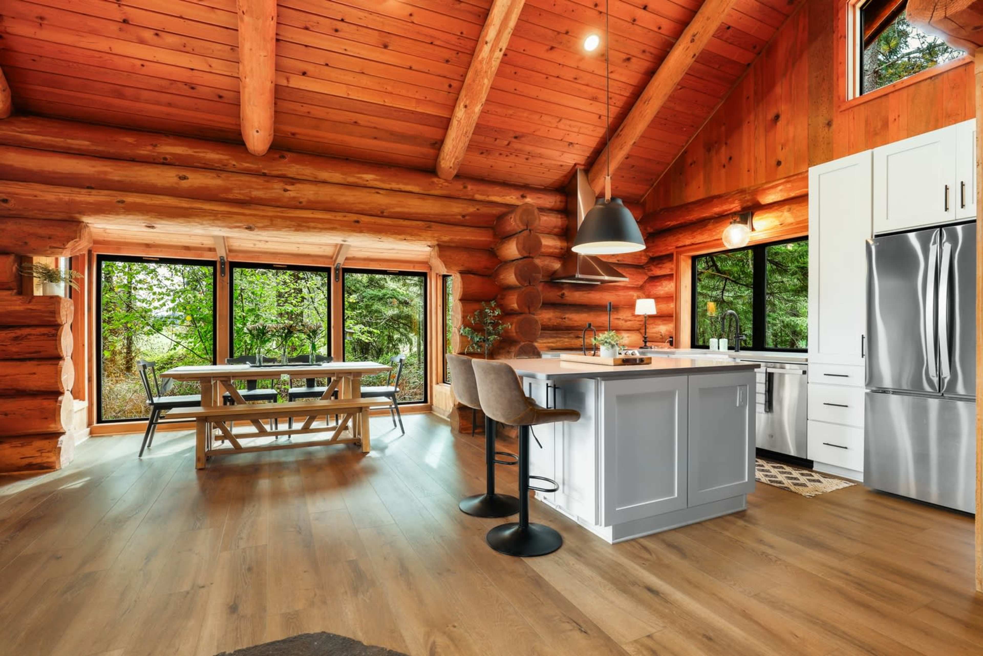 The image shows a spacious log cabin kitchen and dining area featuring wood beams, large windows, a central island, and a dining table.