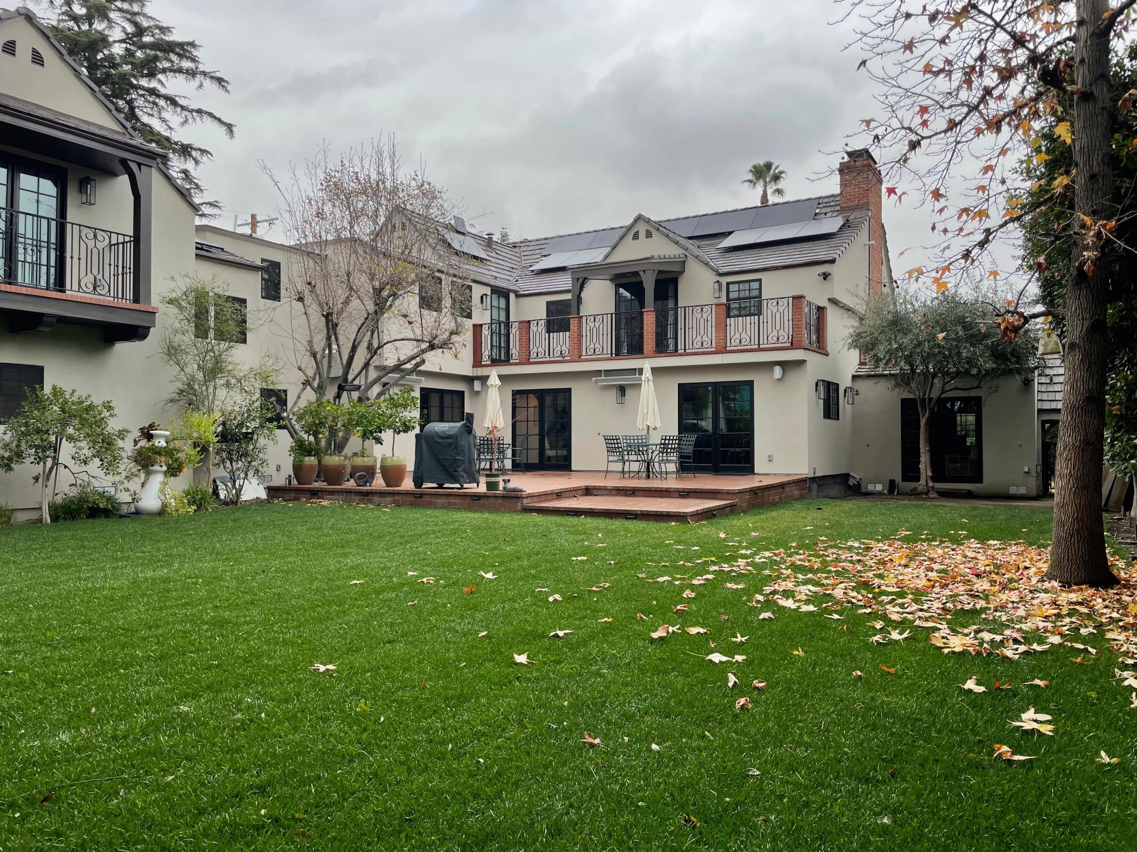 A two-story house with a balcony overlooks a green lawn scattered with fallen leaves.