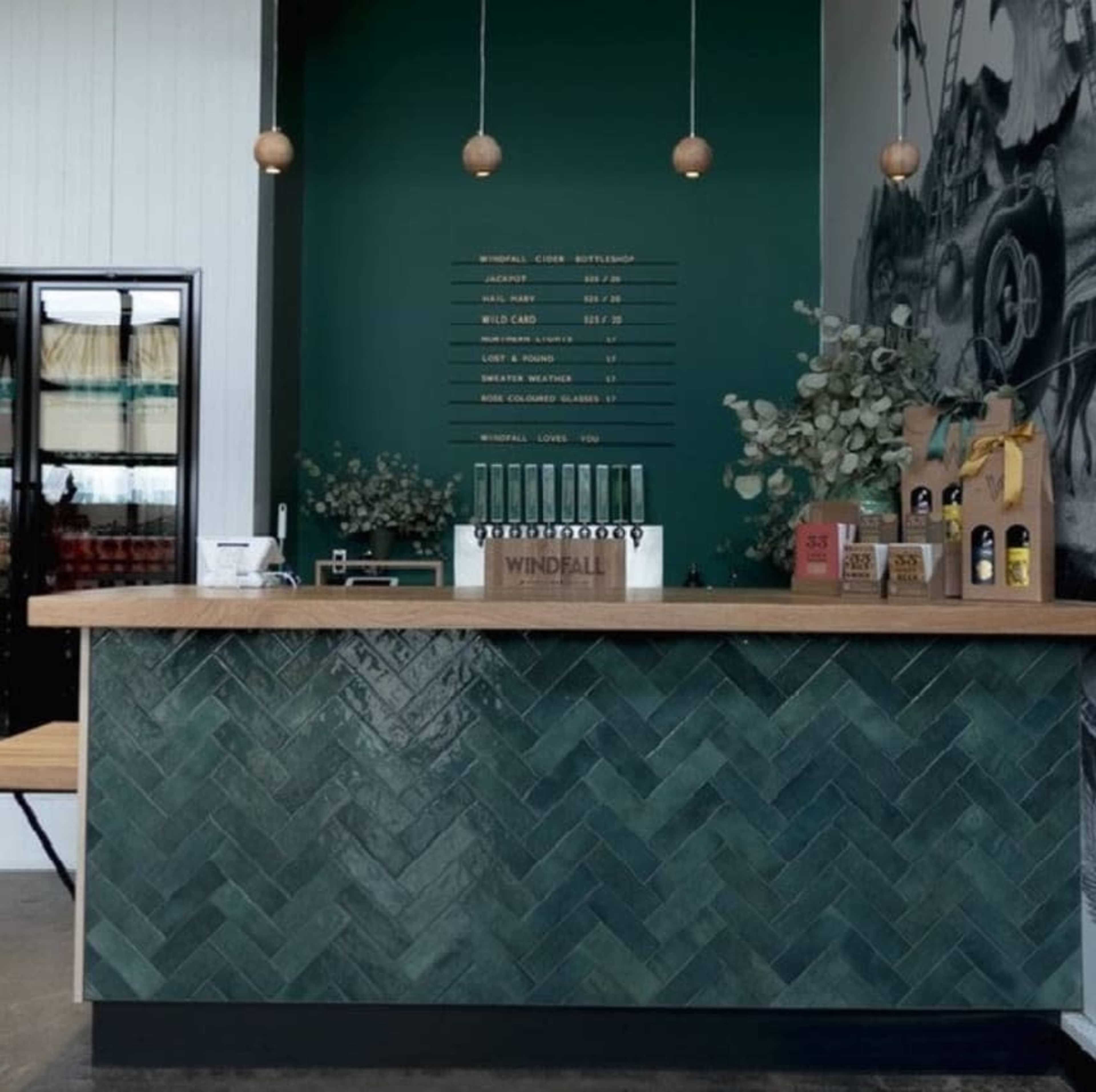The image shows a bar counter with a herringbone-patterned green tile surface, against a dark green wall displaying a menu, with pendant lights hanging above and various plants and products arranged on the counter.