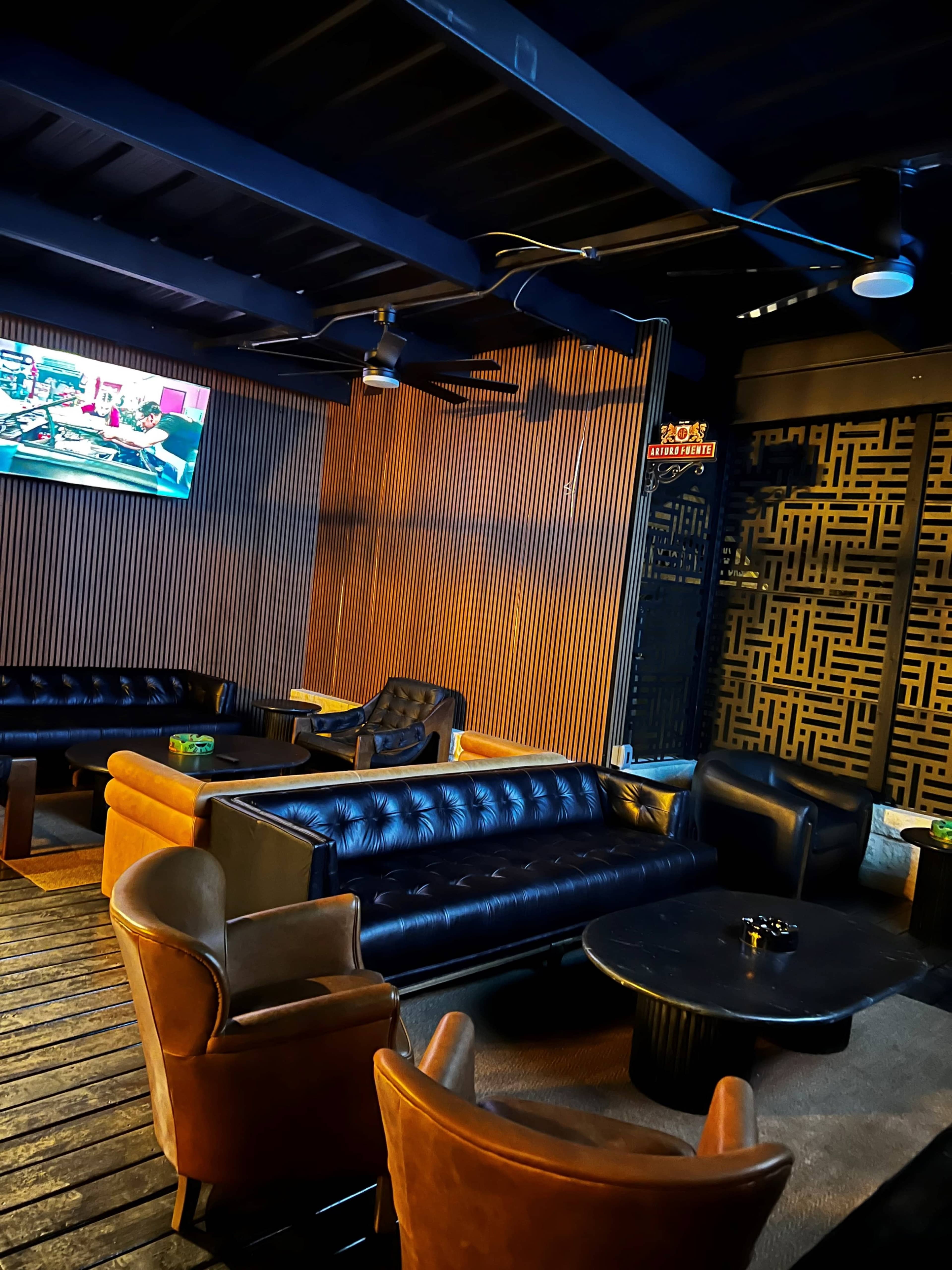 The image shows a dimly lit lounge area with black leather sofas, wooden walls, and a television displaying a show in the background.