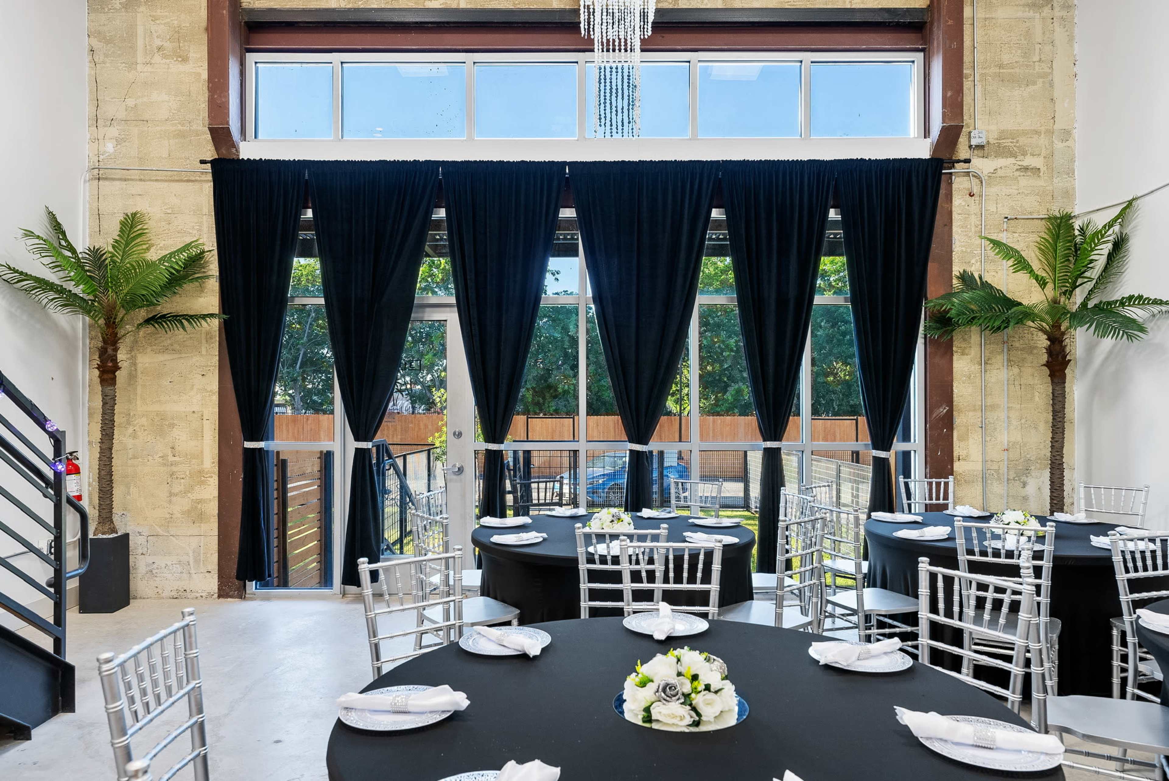 An elegantly set dining area with round tables covered in black cloth, silver chiavari chairs, and large windows draped with black curtains.