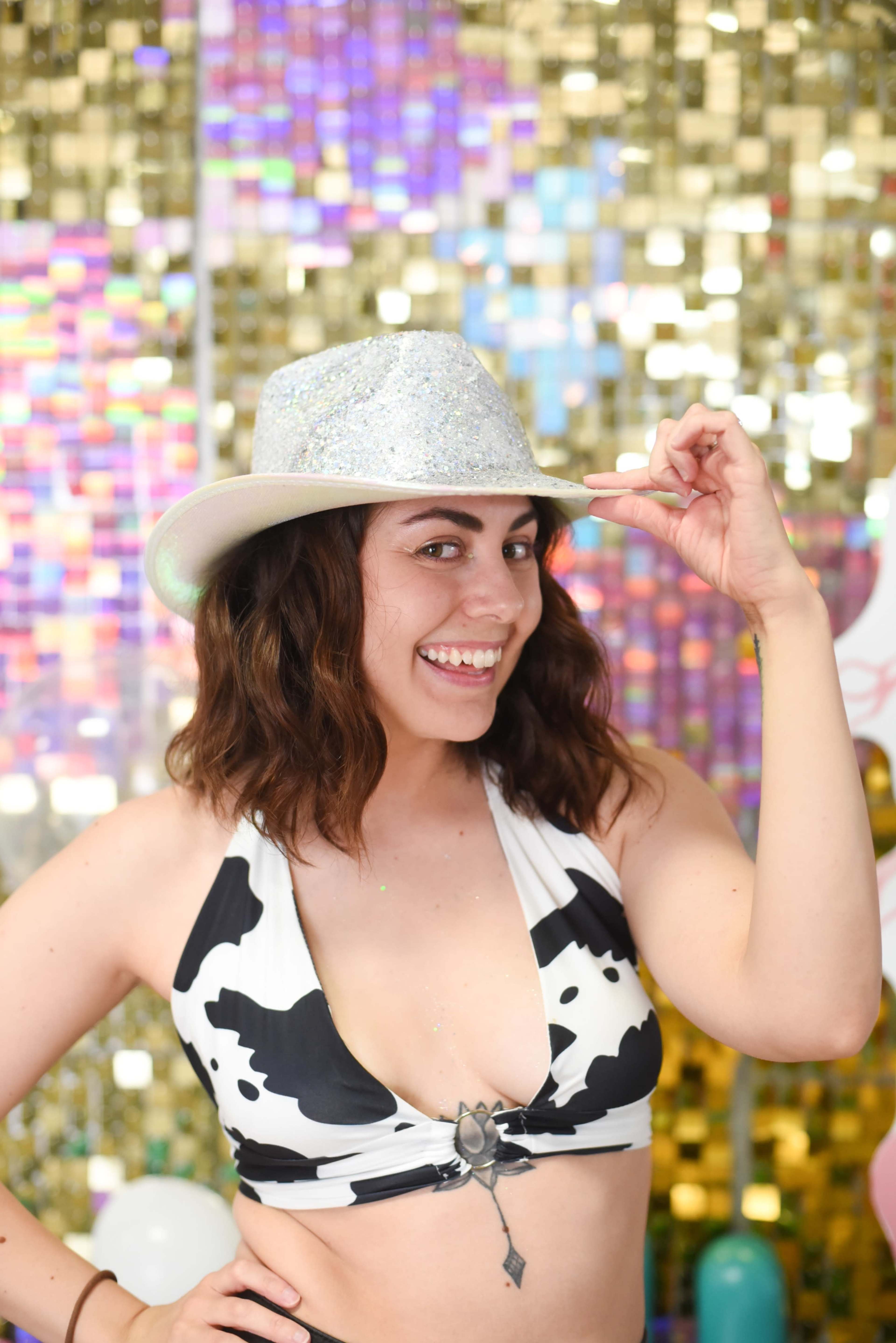 A woman in a cow-print bikini and a sparkling silver hat poses with a smile against a shiny, colorful backdrop.