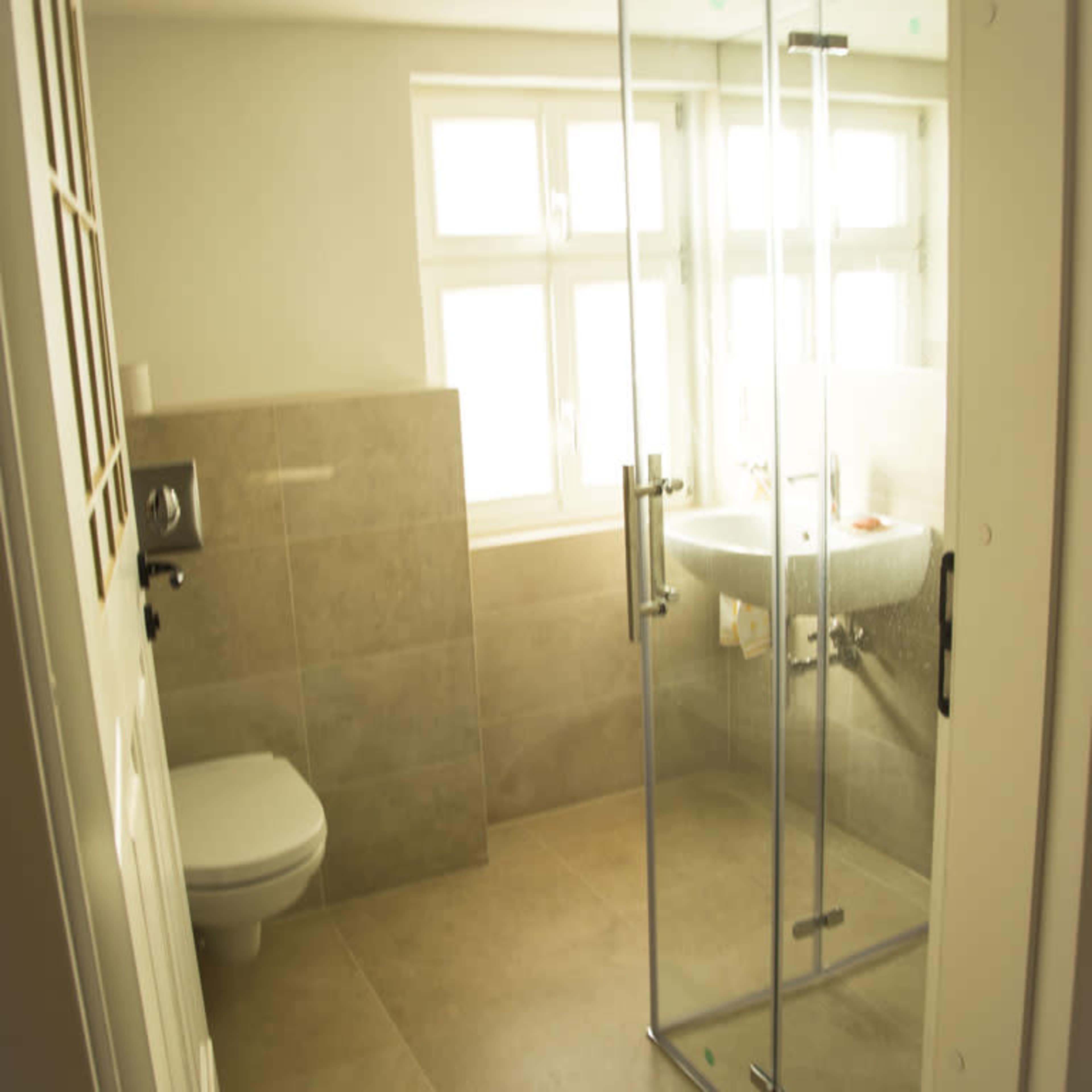 A simple bathroom featuring a glass shower enclosure, a toilet, and a sink next to a row of large windows.