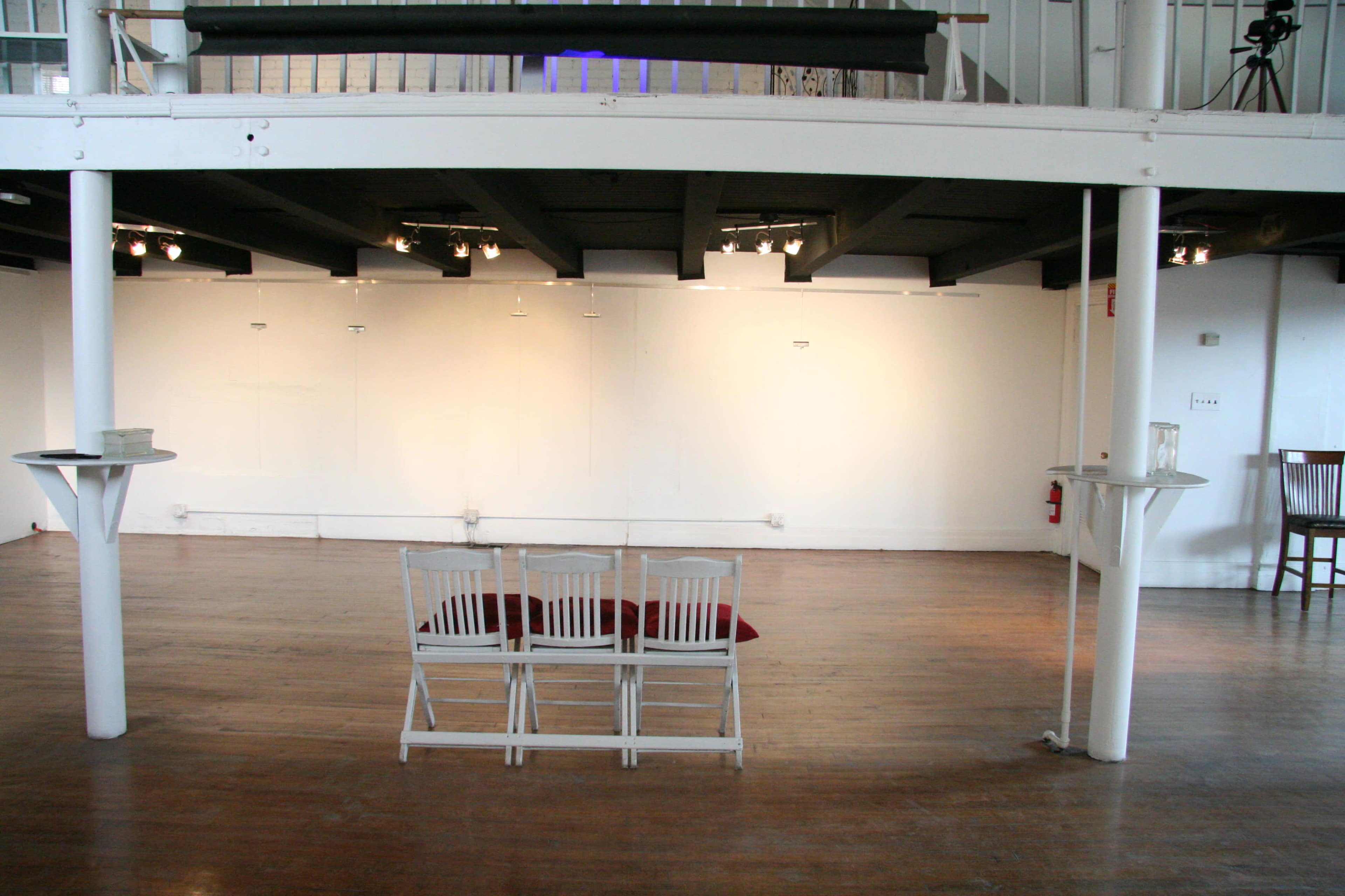 An empty gallery space features three white chairs arranged on a red rug against a blank wall, with wooden floors and exposed beams above.