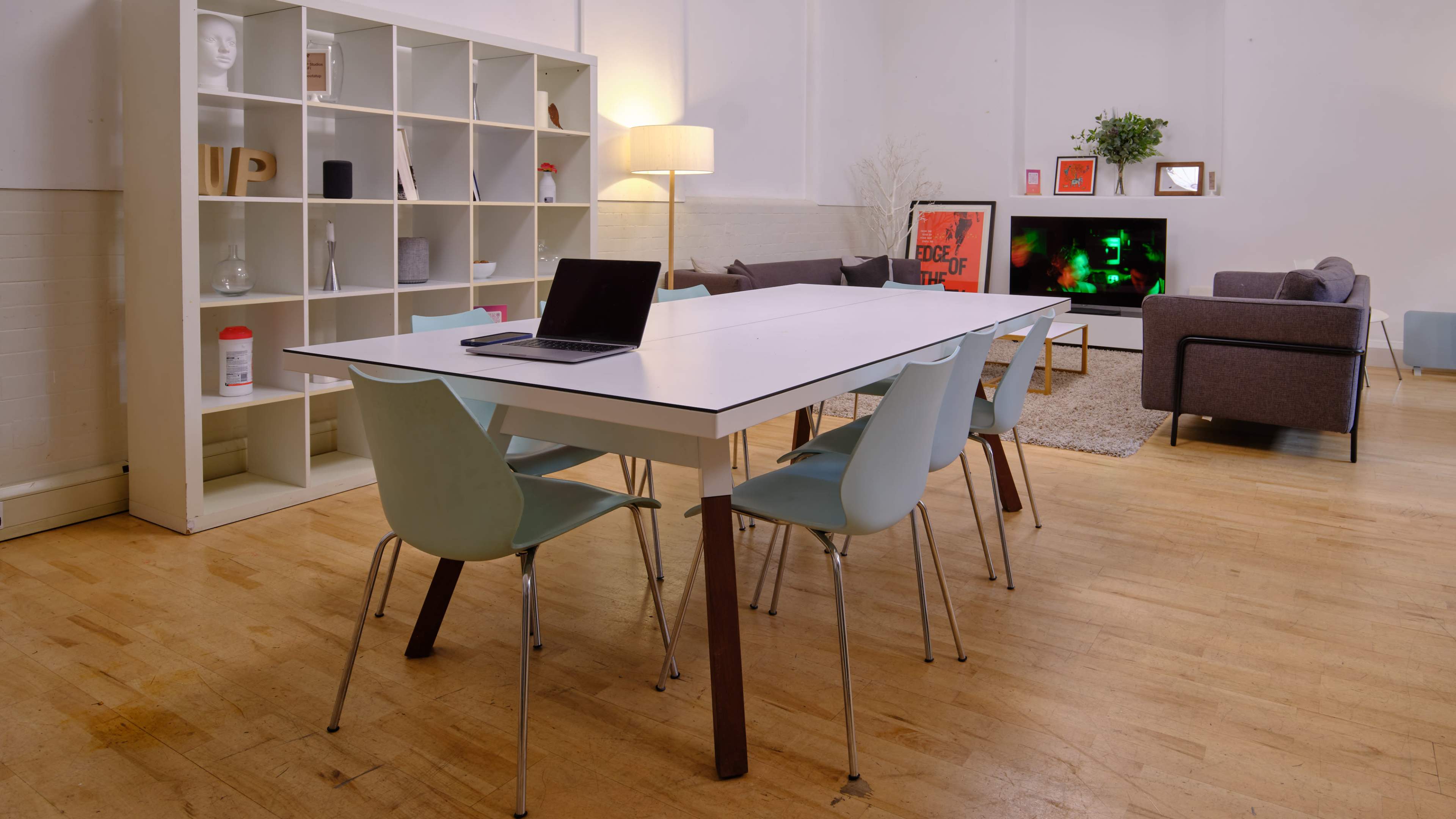 A modern interior features a long white dining table with chairs, a bookshelf, a lamp, and a television in a brightly lit room.