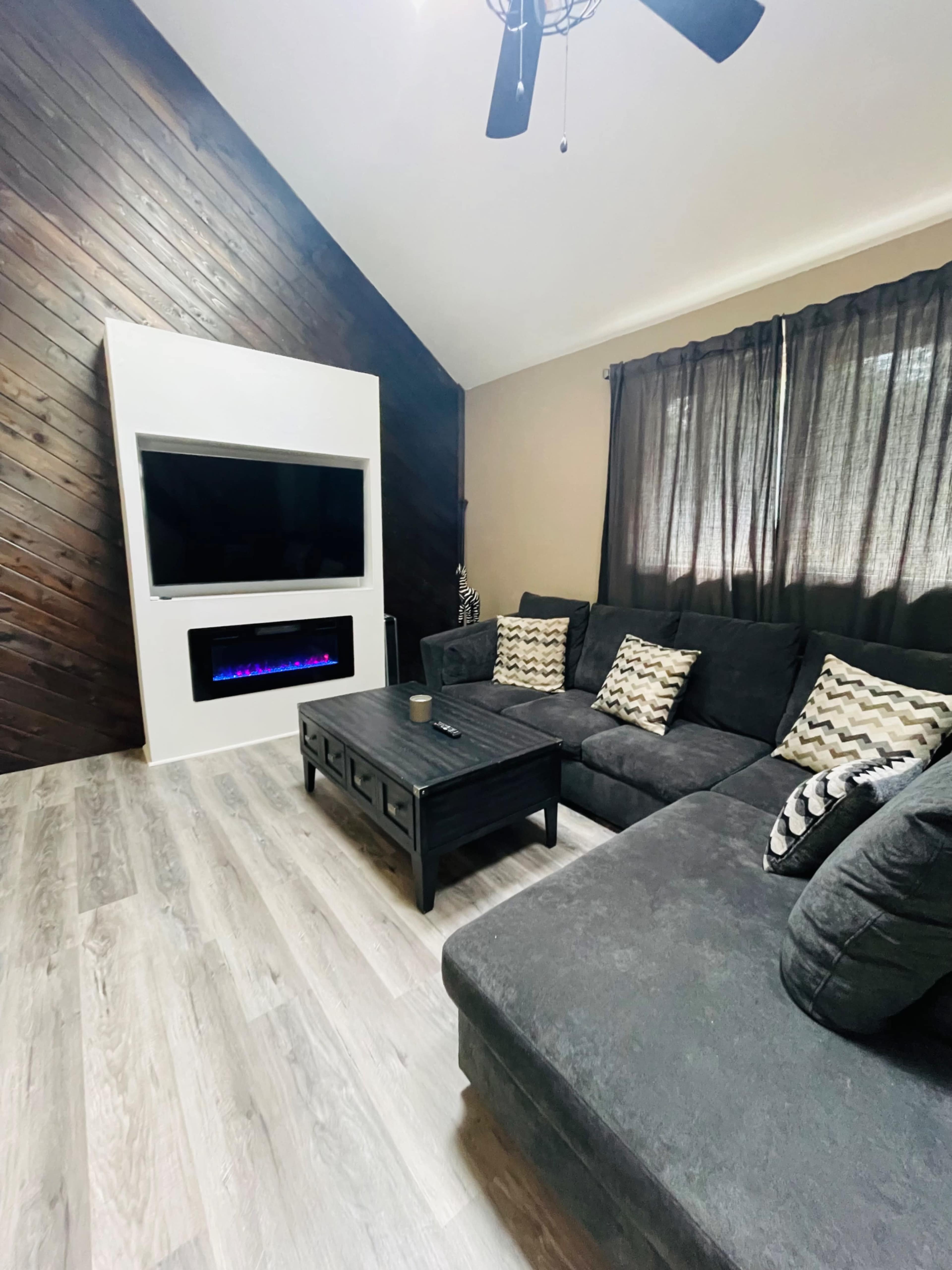A modern living room with a sectional sofa, a coffee table, an electric fireplace beneath a mounted television, and dark curtains on the windows.