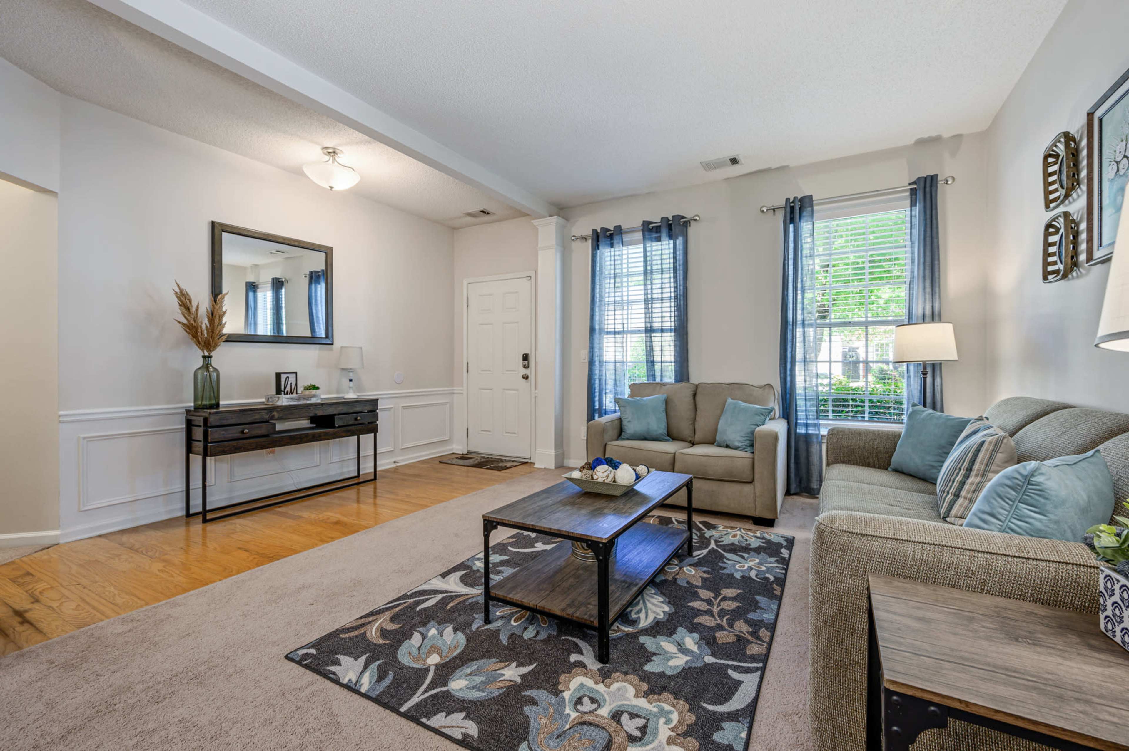 A well-lit living room featuring a sofa, chairs, a coffee table, and decorative accents, with a front door and windows letting in natural light.