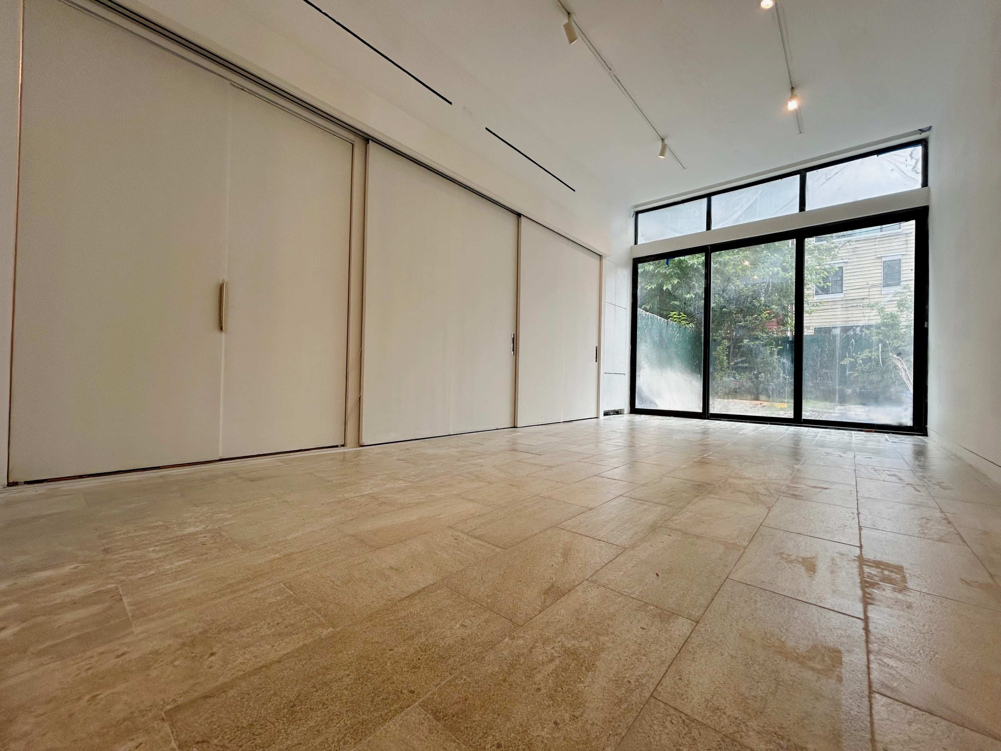The image shows an empty room with large glass doors opening to a garden, and a light-colored tiled floor.