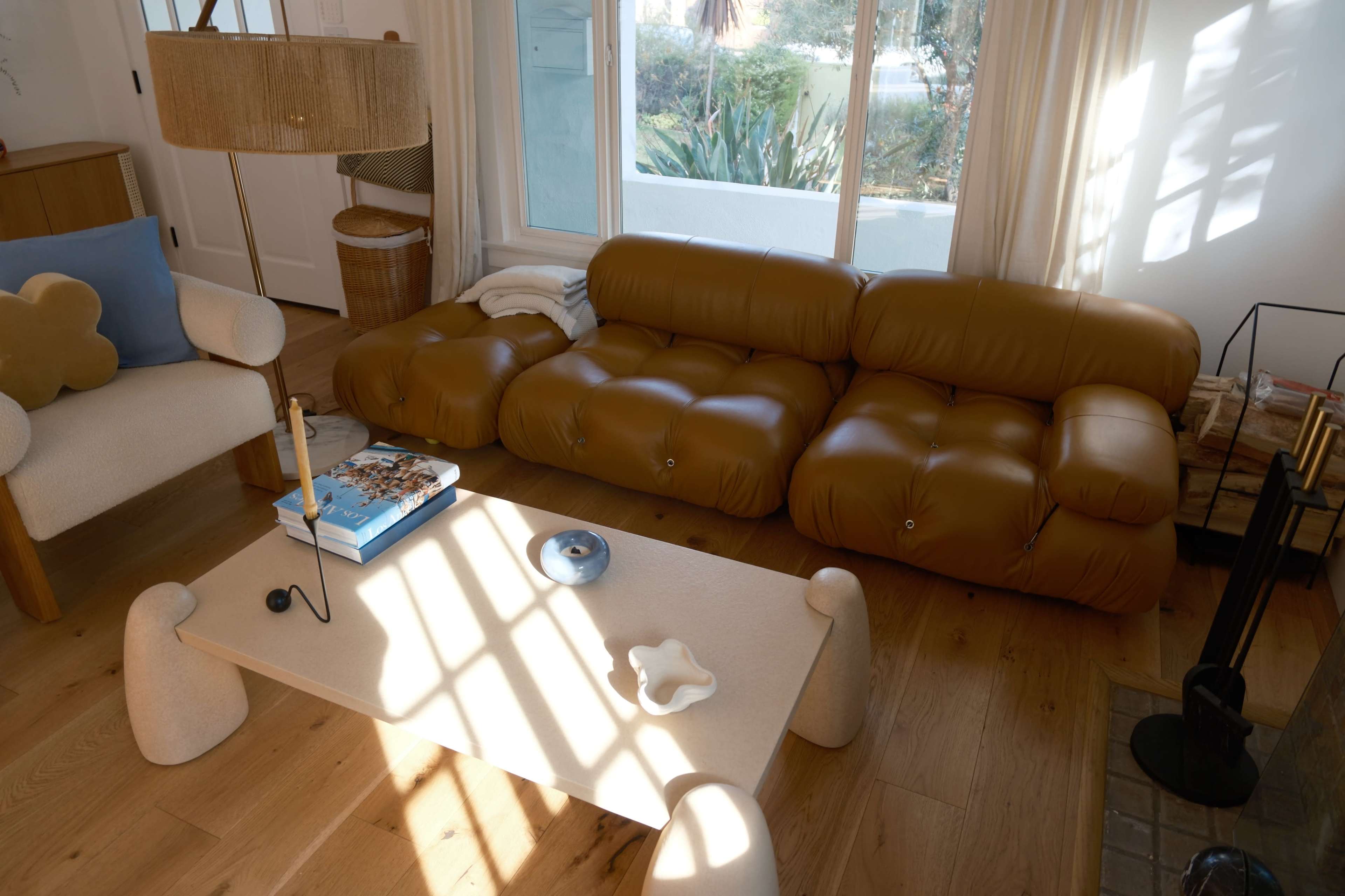 The scene features a modern living room with a brown, cushioned sofa, a light-colored coffee table, and large windows allowing natural light to enter.
