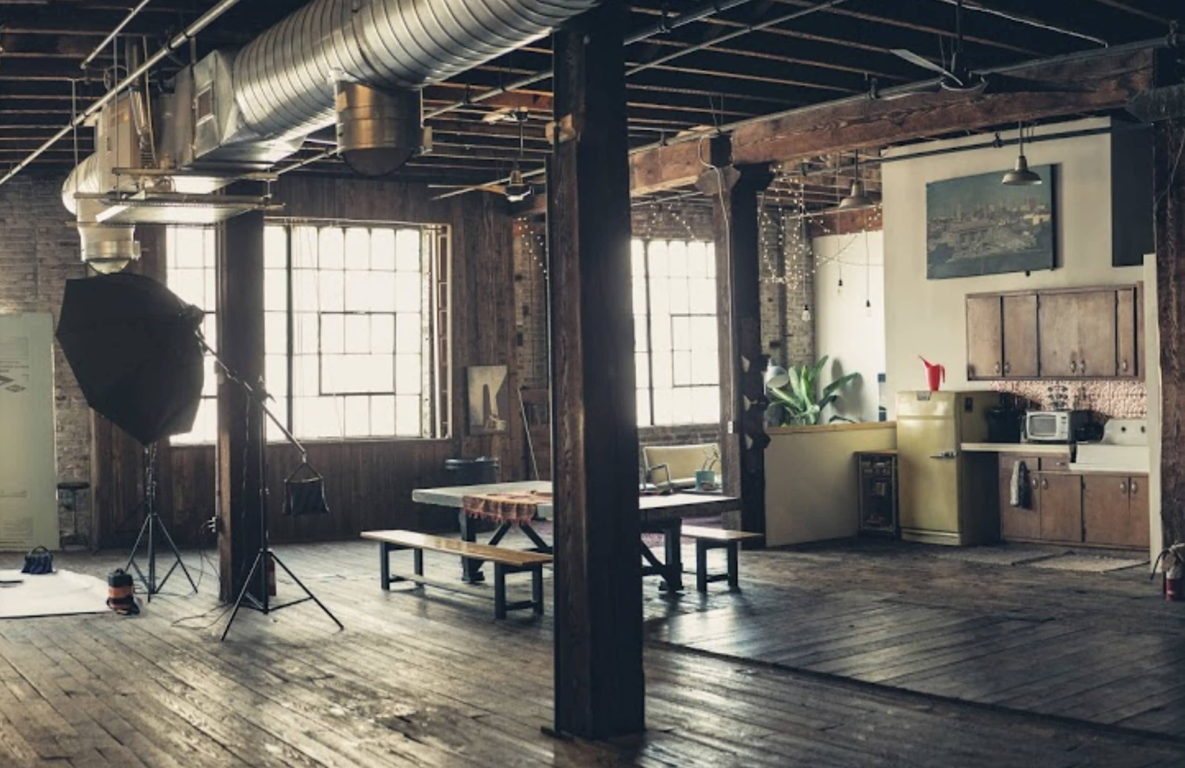 The image shows a spacious industrial-style room with wooden beams, large windows, and a mix of vintage and modern furniture, including a yellow fridge and a photography setup.