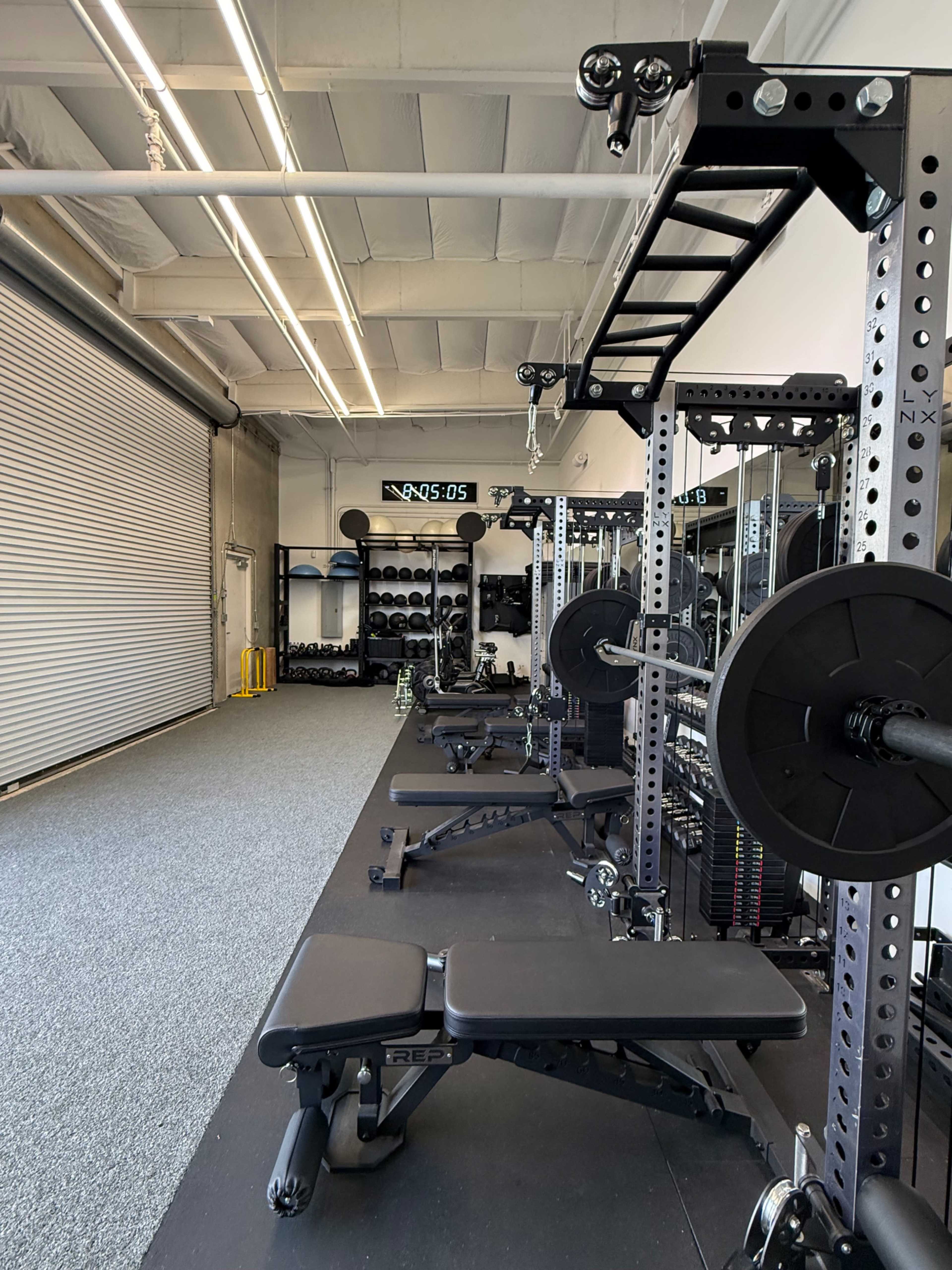 The image shows a well-equipped gym space with weightlifting benches and a power rack, lined along a wall with shelves of weights in the background.