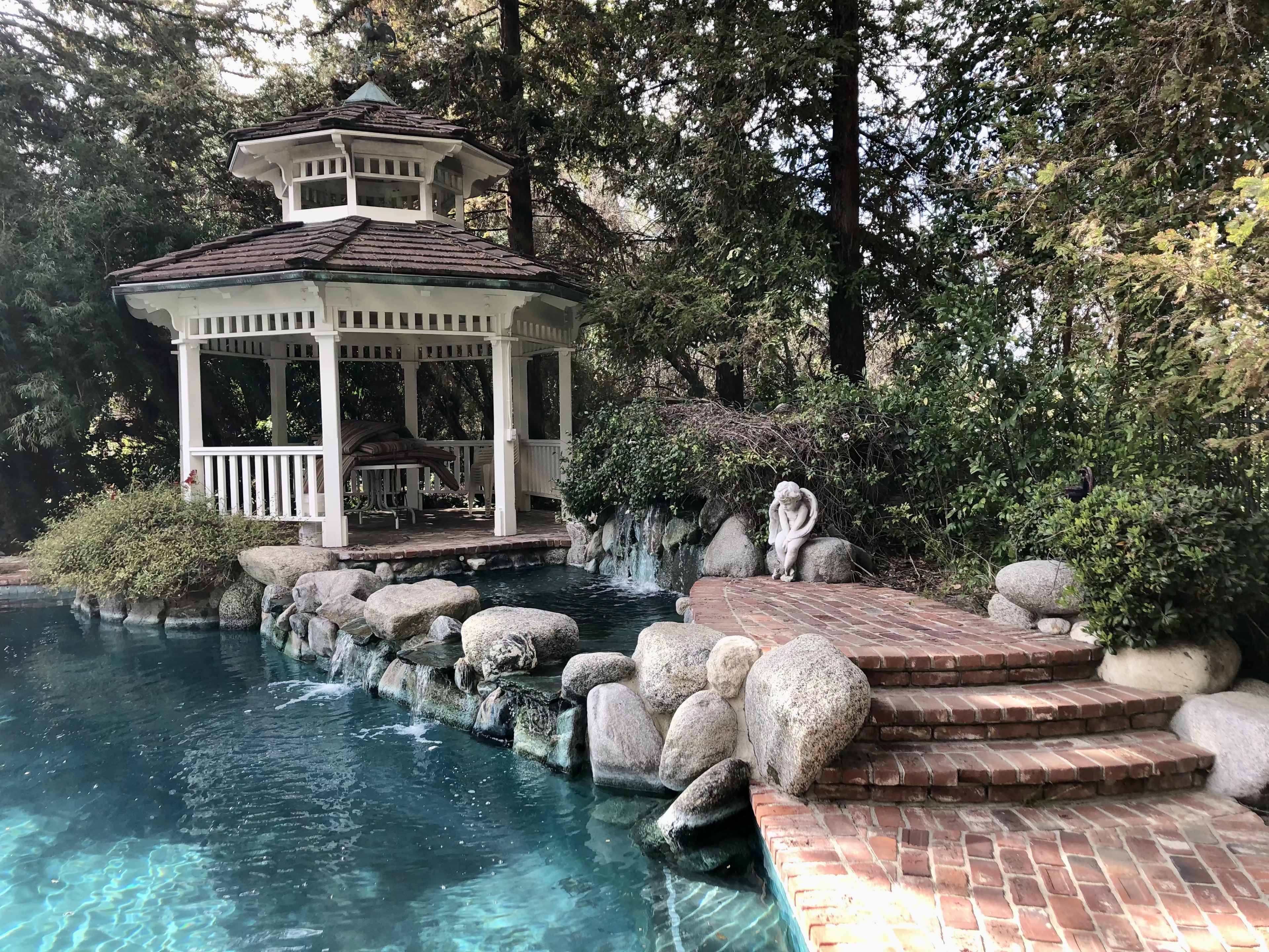 A gazebo with a shingled roof stands near a pool surrounded by stone steps and lush greenery.