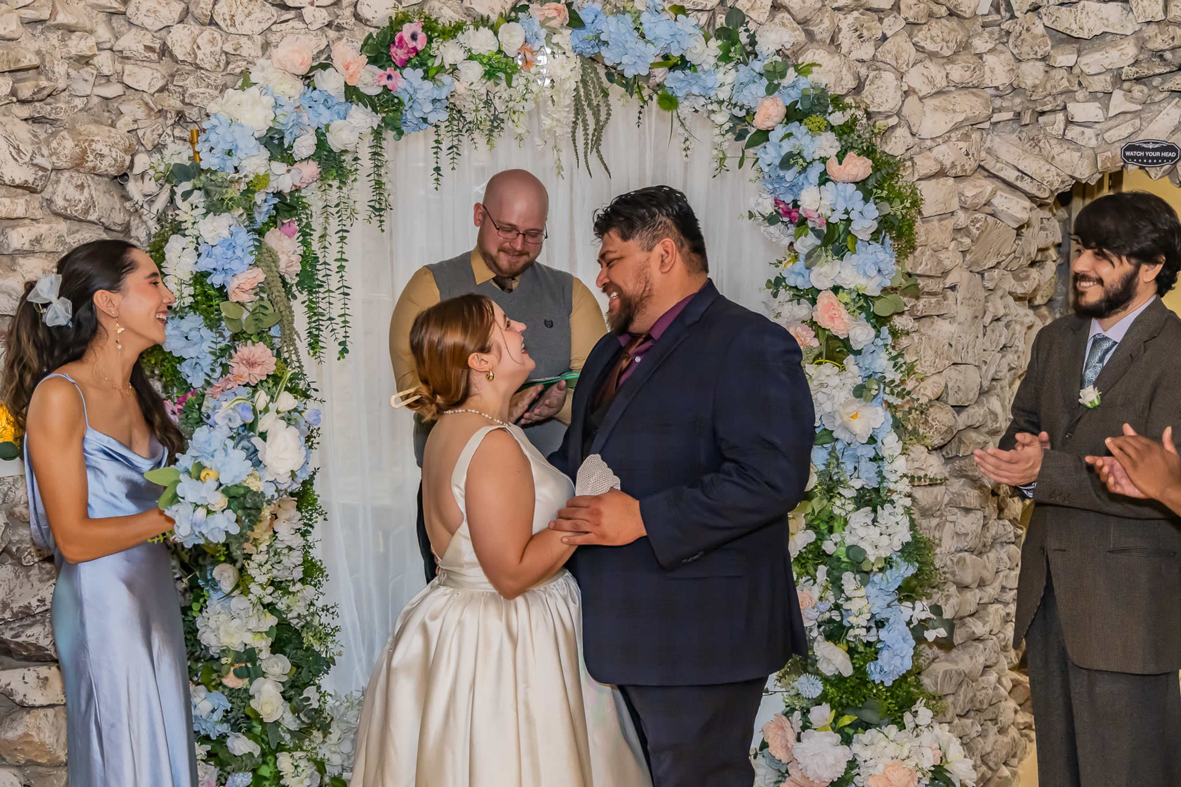 A couple stands in front of a floral arch during a wedding ceremony while a person officiates in the background.