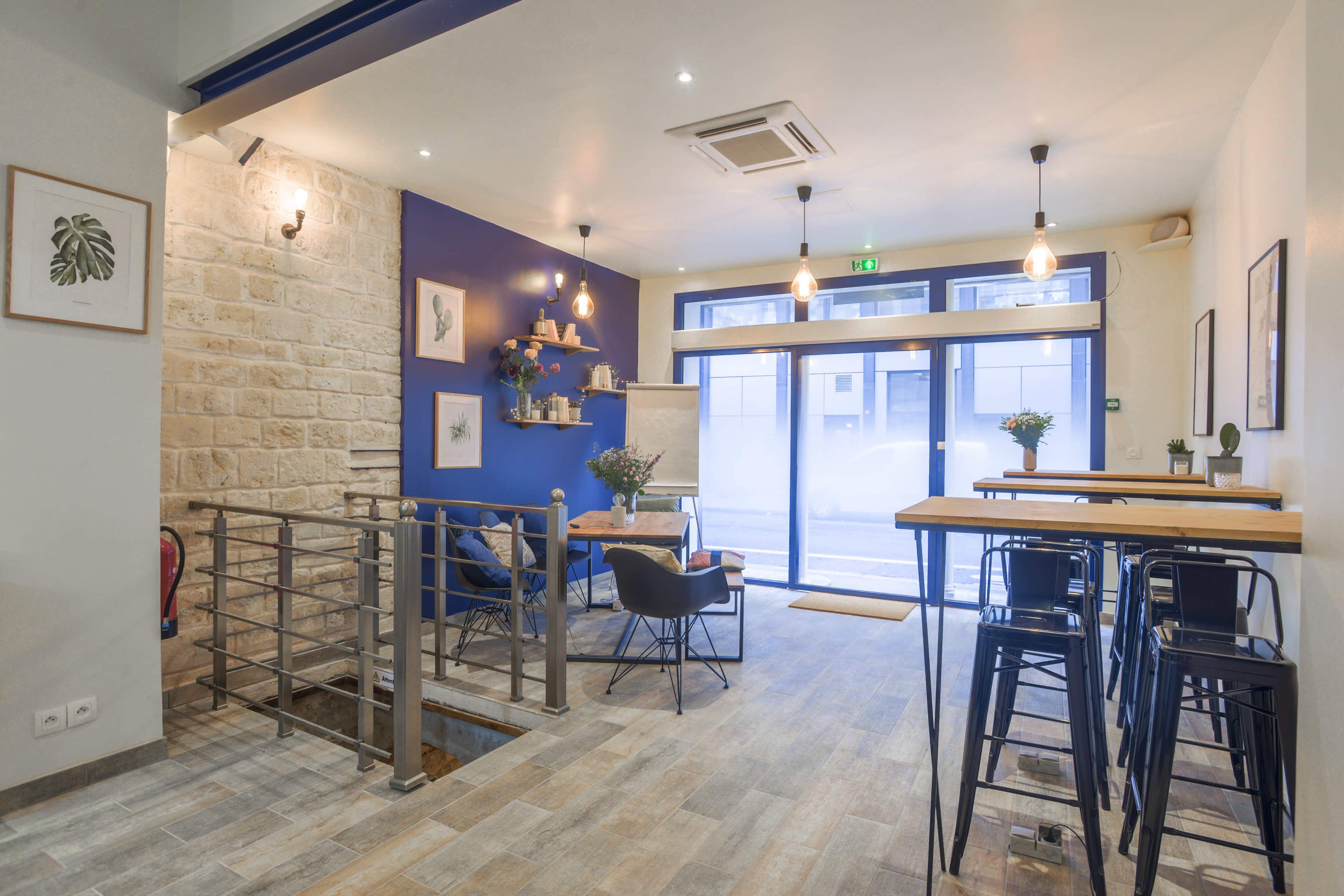 A modern café with a light-colored stone wall, wooden flooring, and tall tables arranged near a large window.