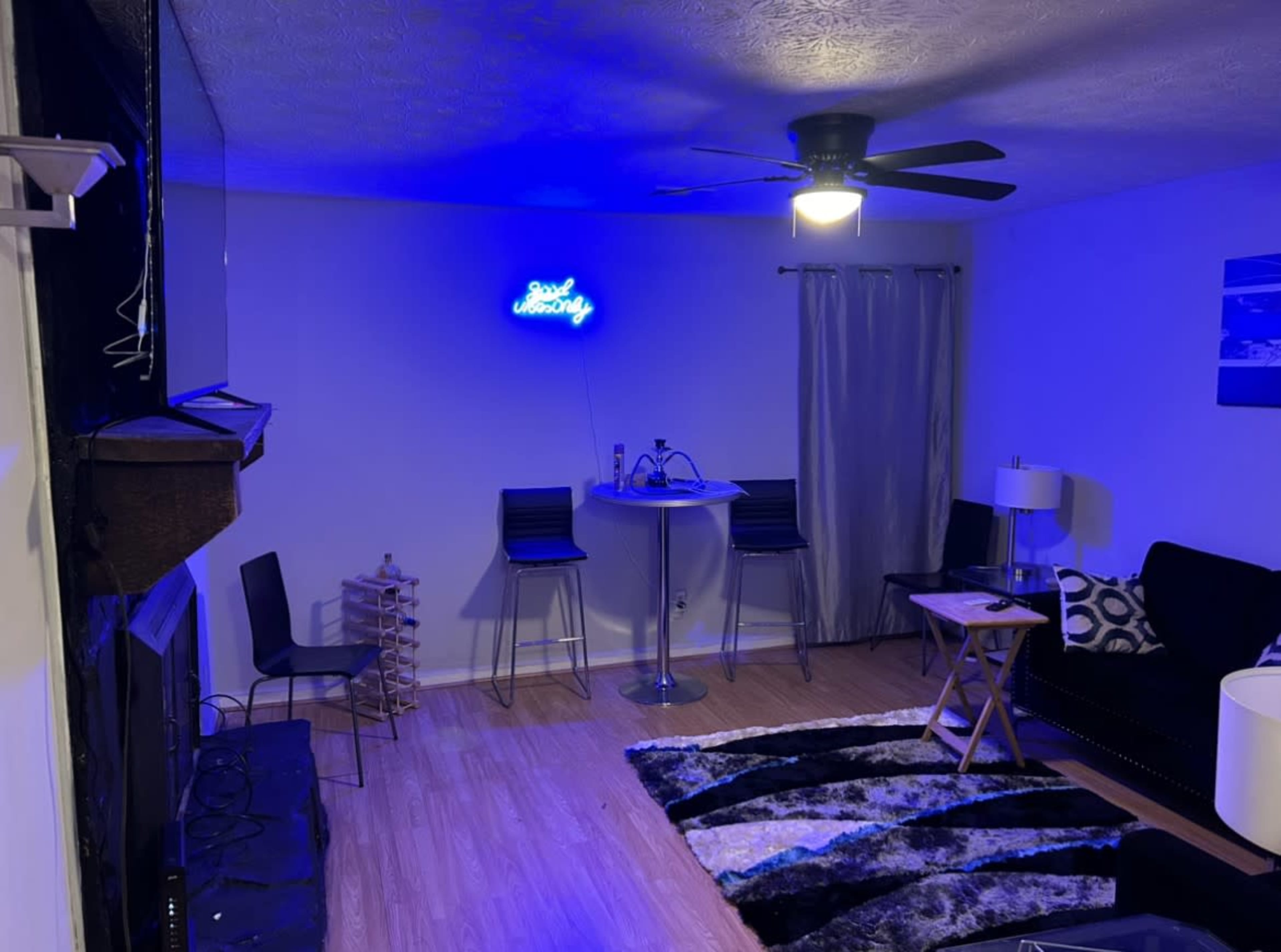 A modern living room features a black couch, a round bar table with stools, a wall-mounted television, and a neon sign against a light-colored wall.