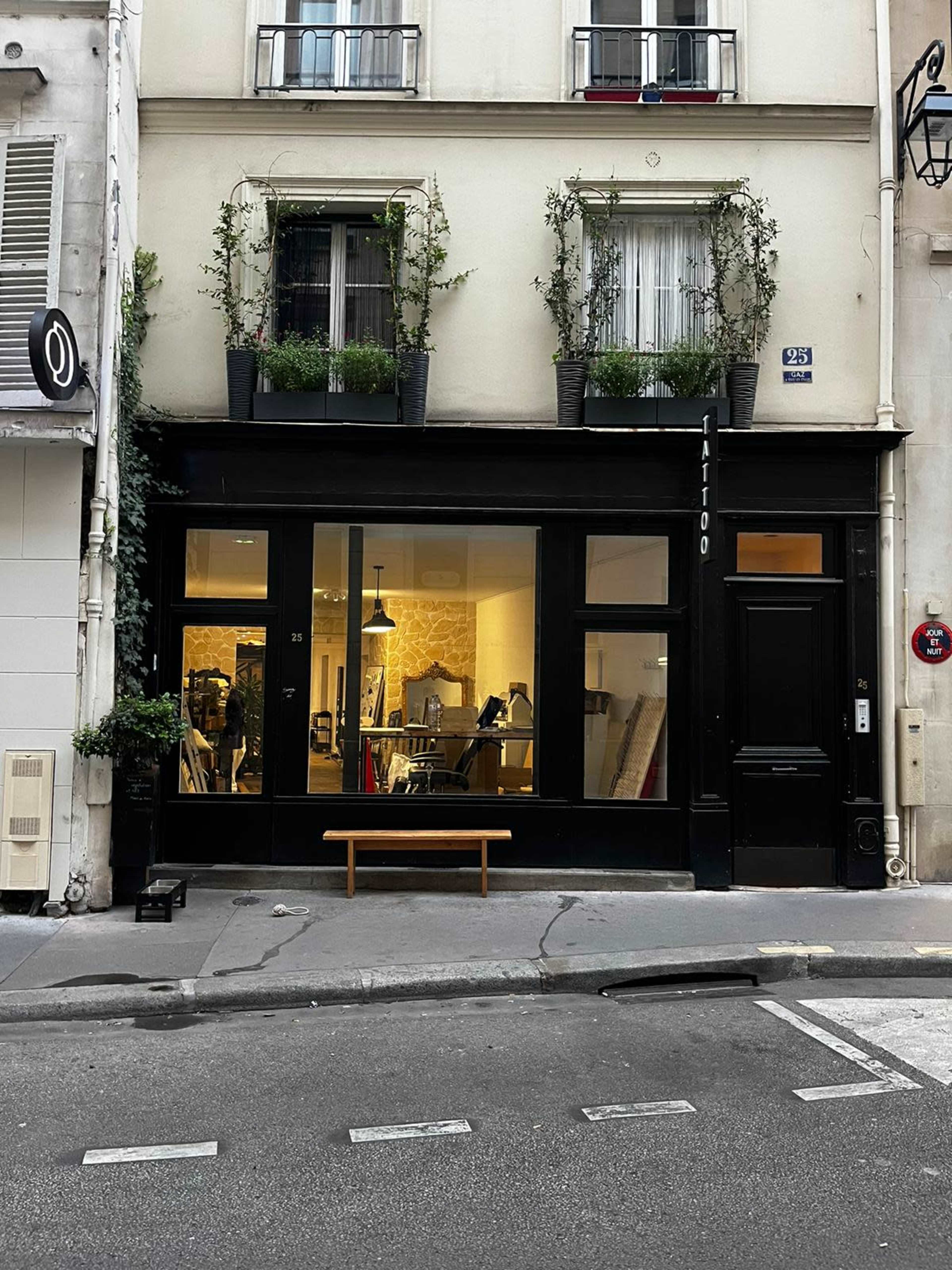 A black storefront with large windows displaying a tattoo studio, flanked by plants in window boxes, is situated on a quiet street in Paris.