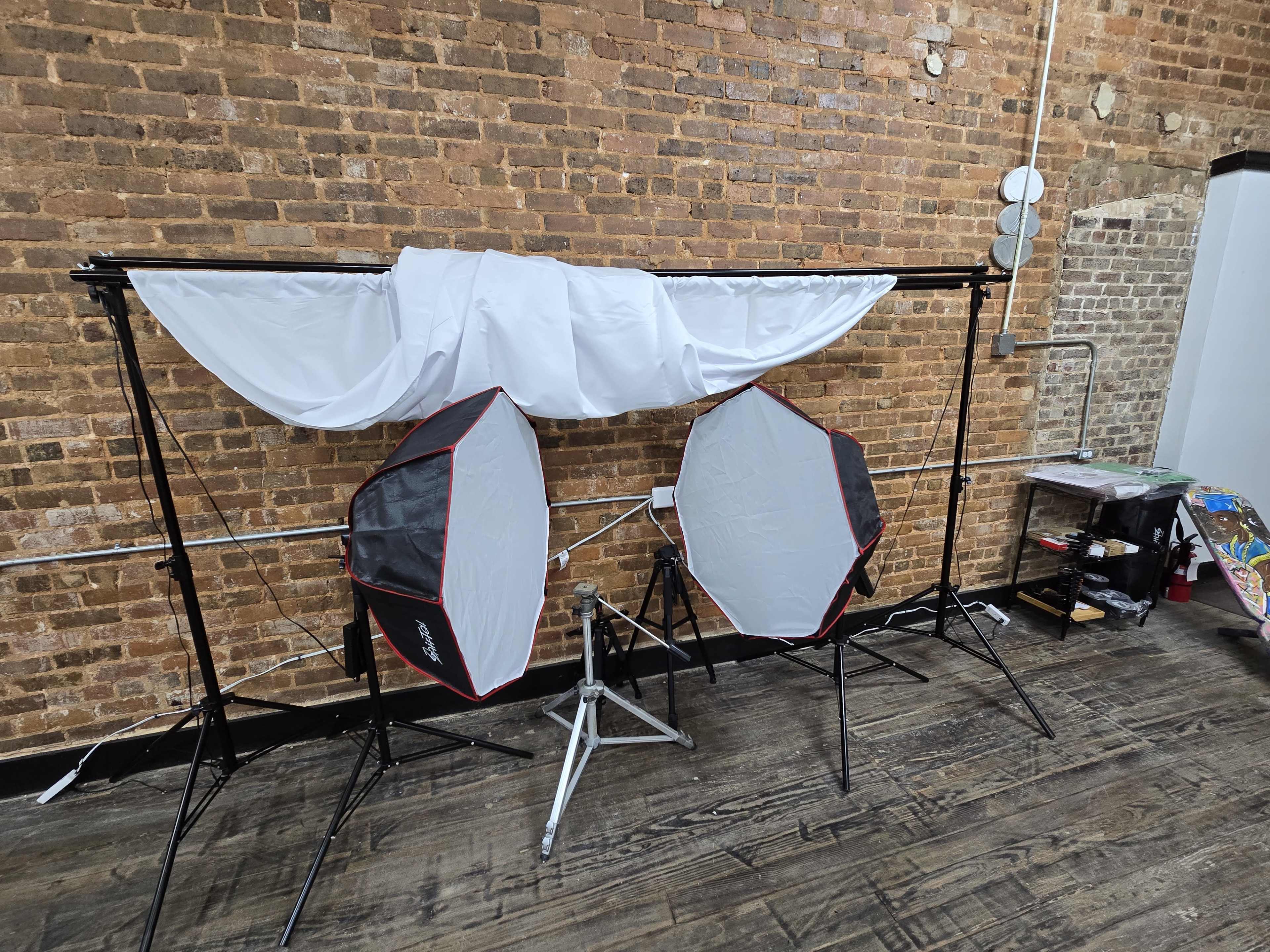 The image shows a photography setup with two softbox lights positioned in front of a draped white backdrop against a brick wall.