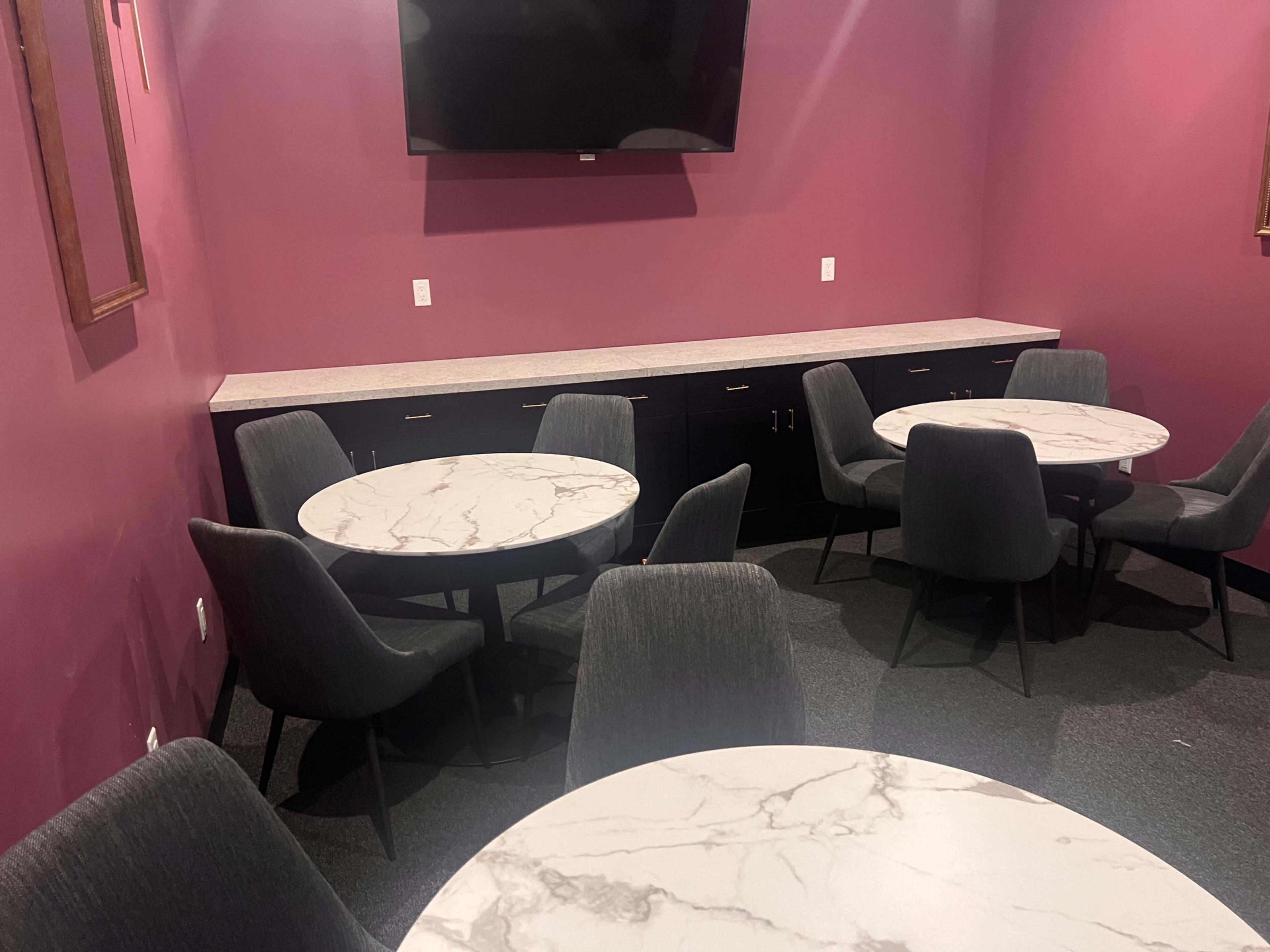 A small room with several round marble-top tables and gray chairs, accompanied by a wall-mounted television and purple-painted walls.