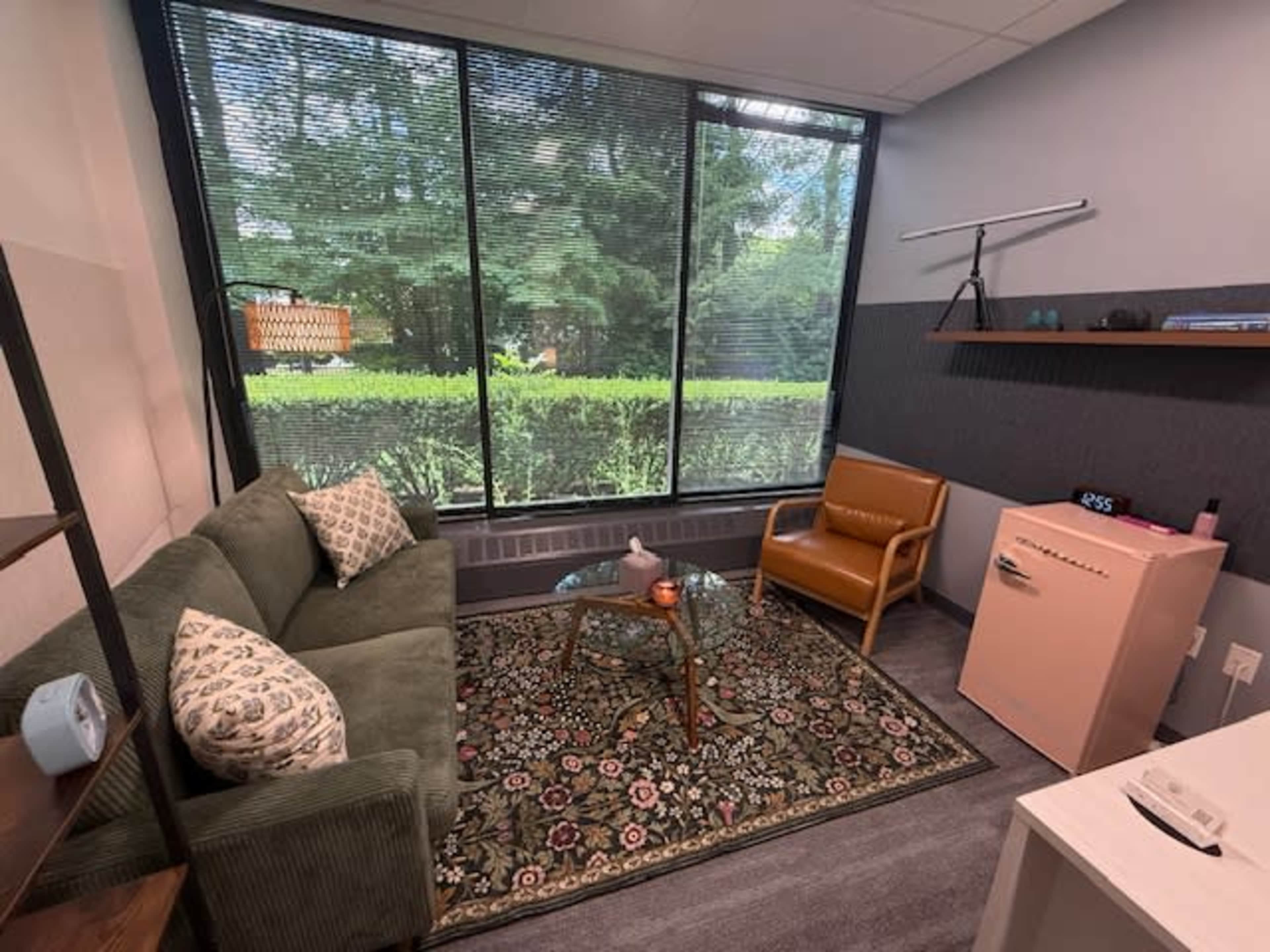 The image shows a cozy office space with a couch, an armchair, a glass coffee table, and large windows displaying greenery outside.