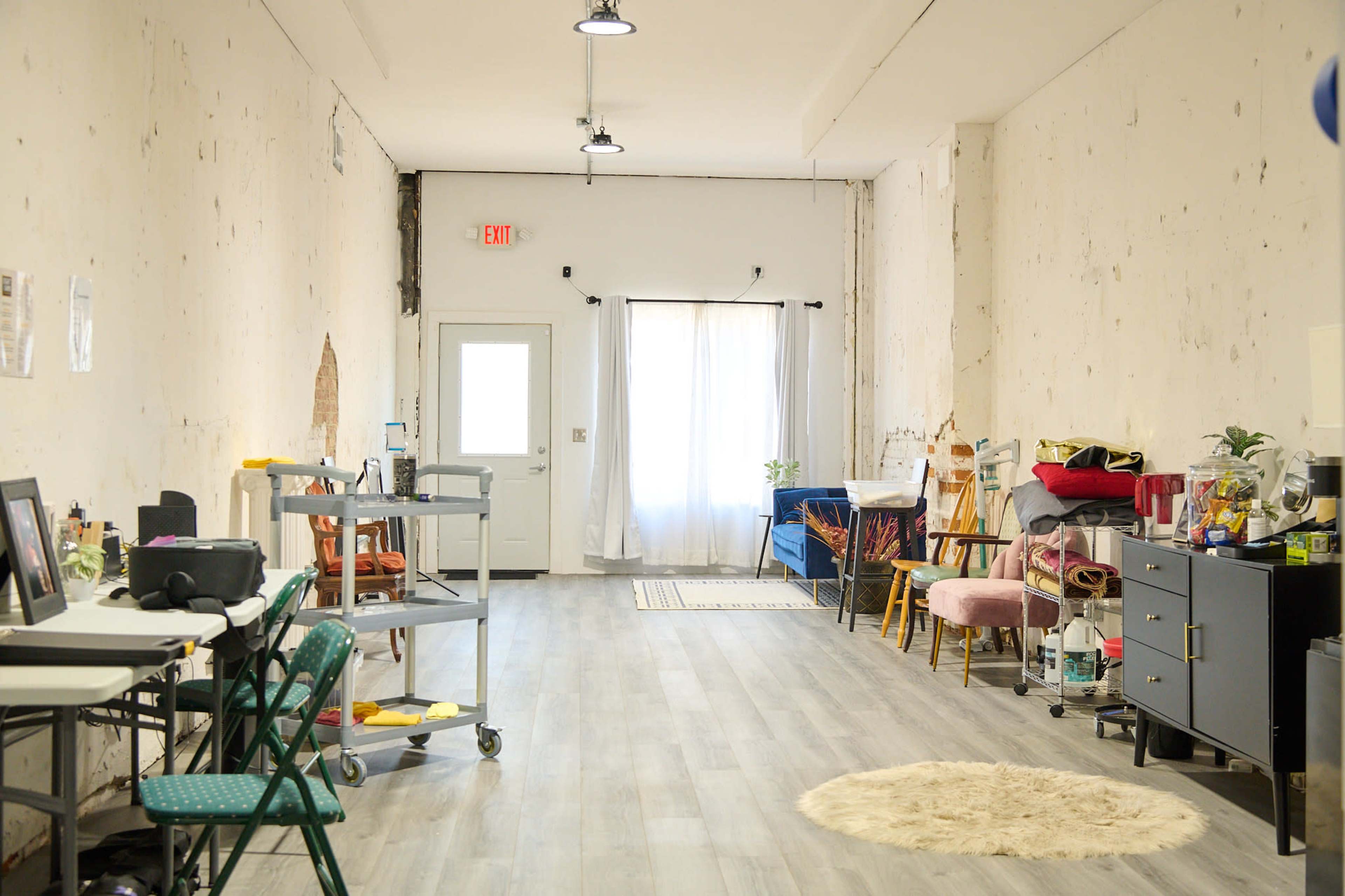 A spacious, minimally furnished room with light-colored walls, a window letting in natural light, and various pieces of furniture along the sides.