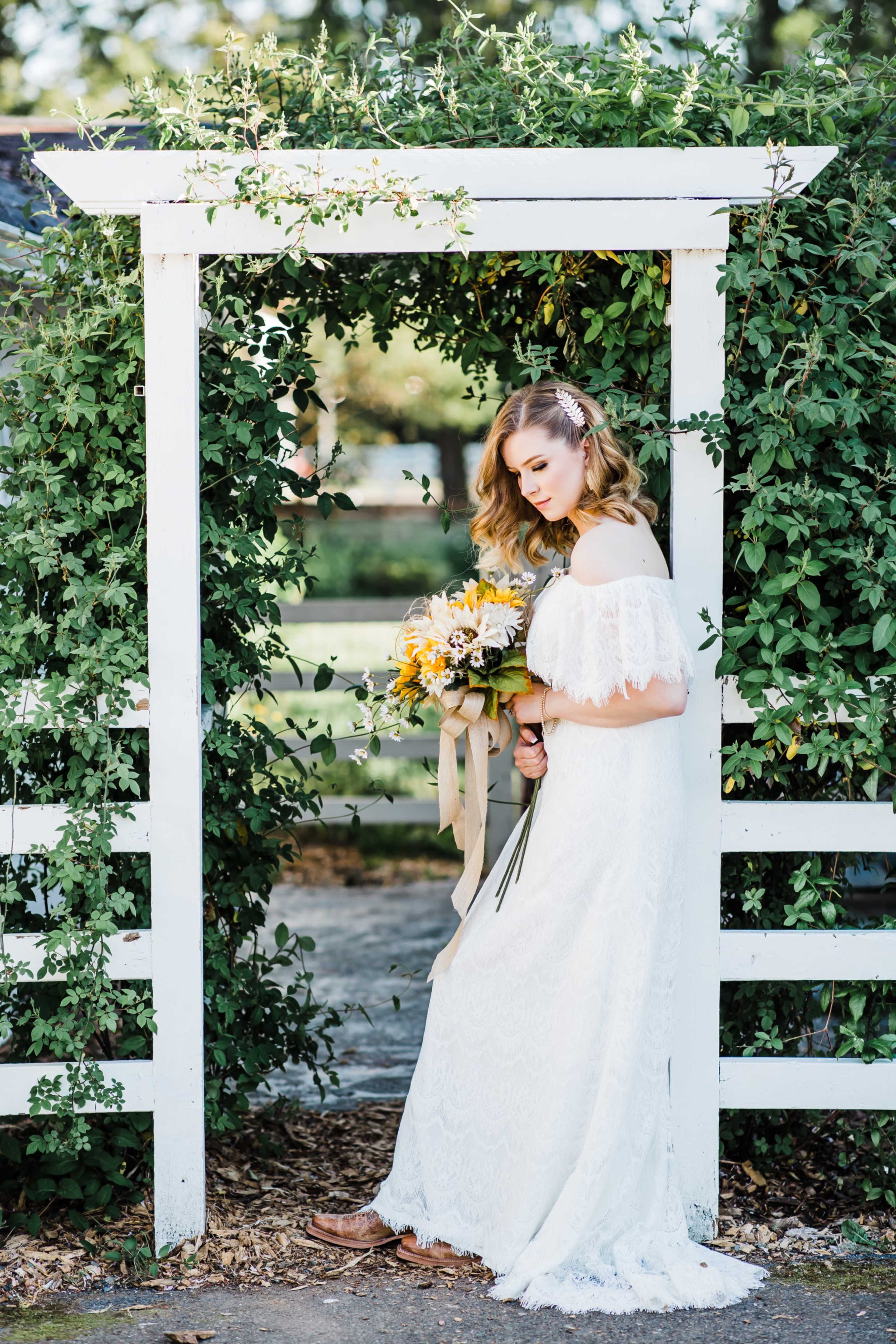 A bride in a white lace gown stands under a green archway adorned with vines, holding a bouquet of flowers.