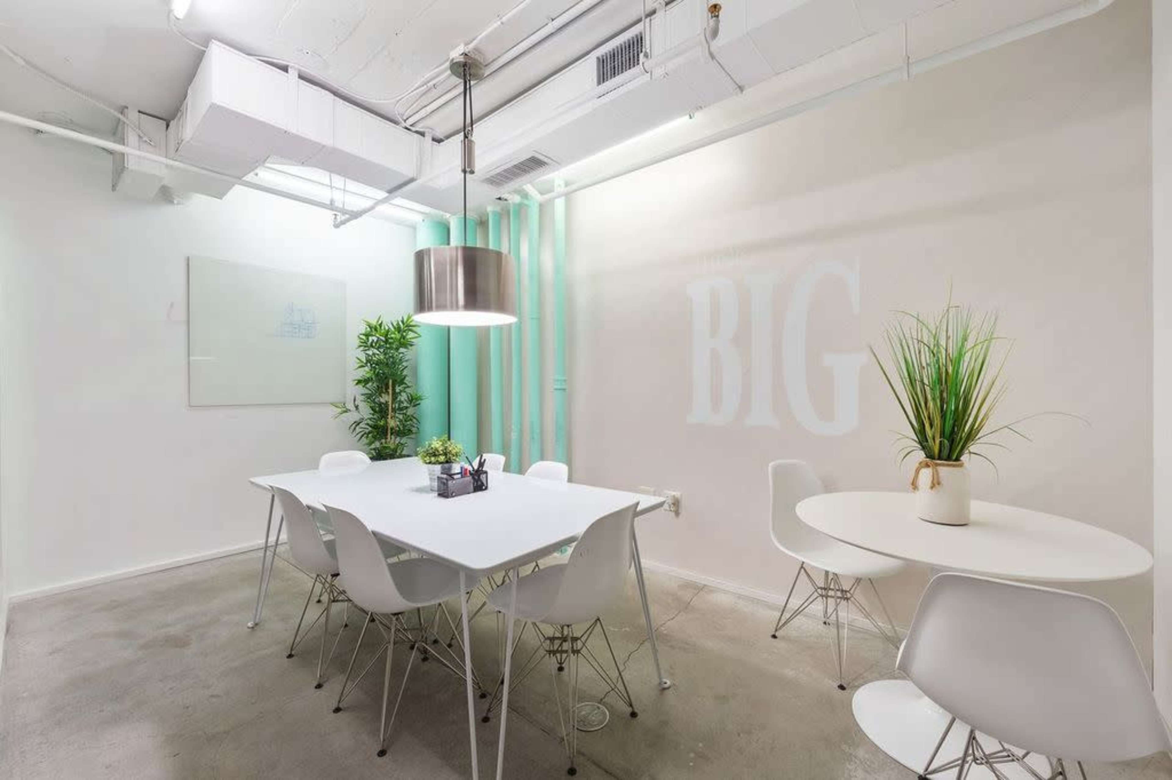 A well-lit meeting room features a white table surrounded by chairs, along with plants and a large pendant light.