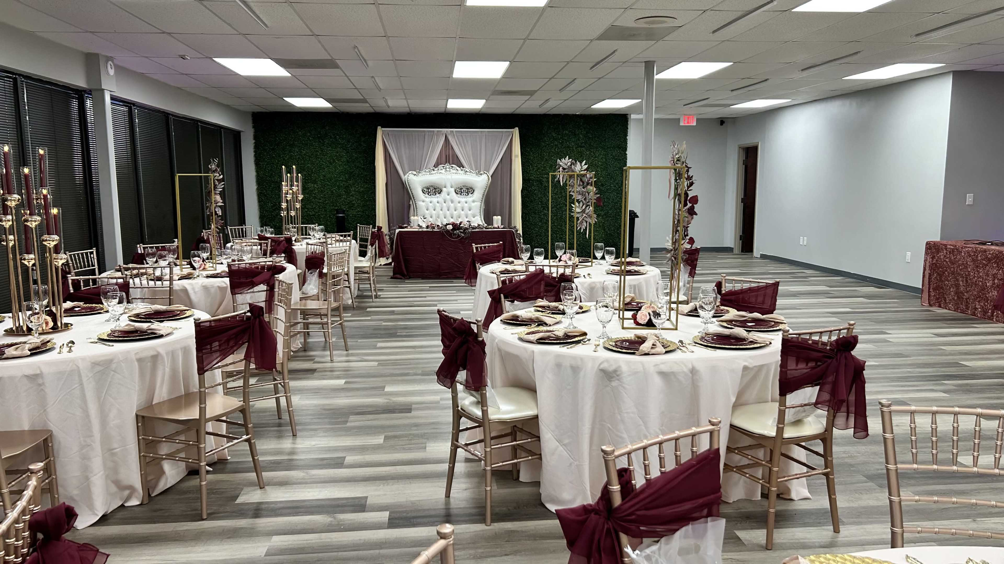 The image shows a banquet hall decorated with round tables dressed in white tablecloths and maroon accents, surrounded by greenery and a backdrop featuring a decorative display at the front.