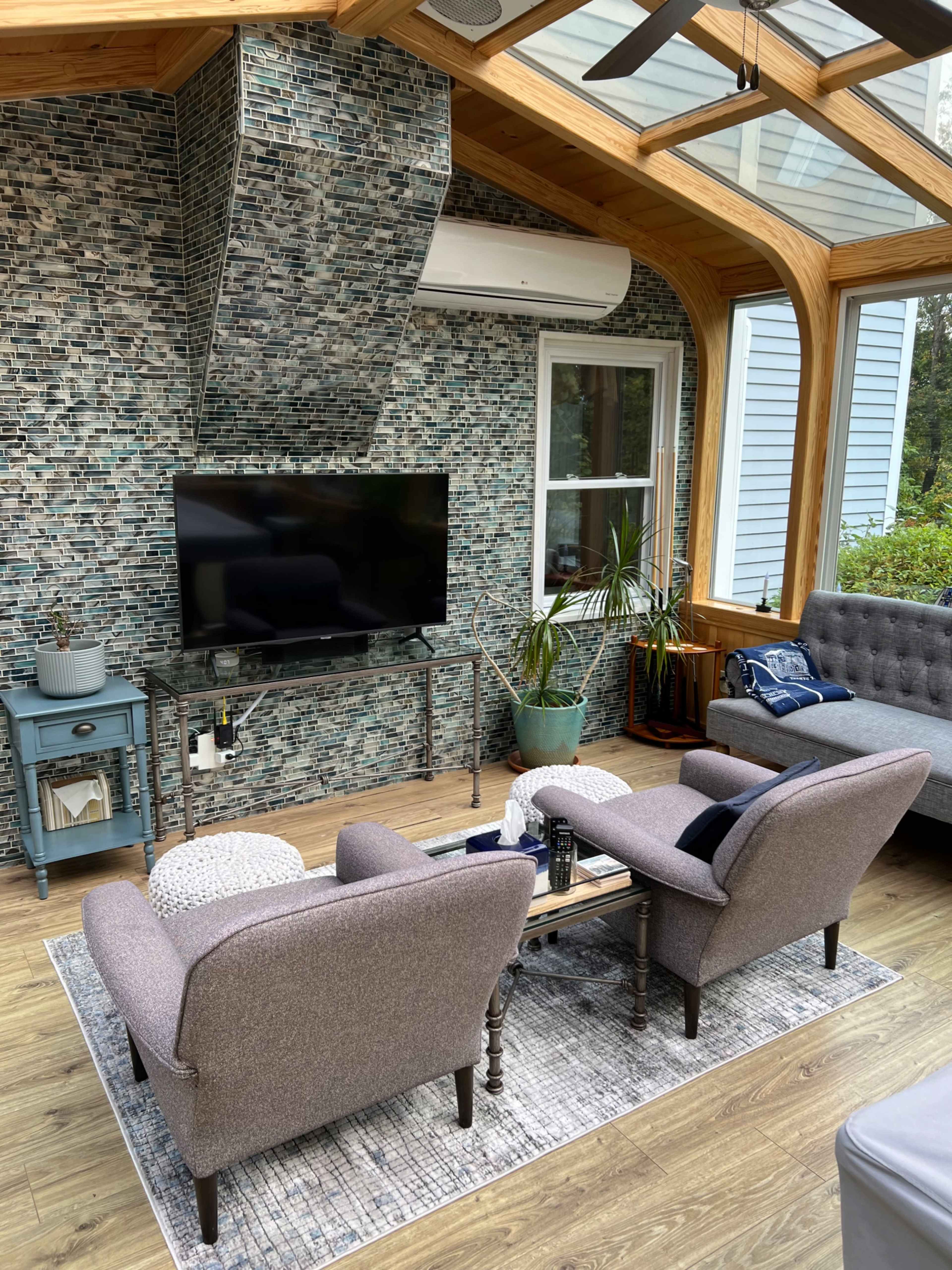 A sunroom features two gray armchairs facing a coffee table, a television mounted on a patterned wall, and a large window overlooking greenery.