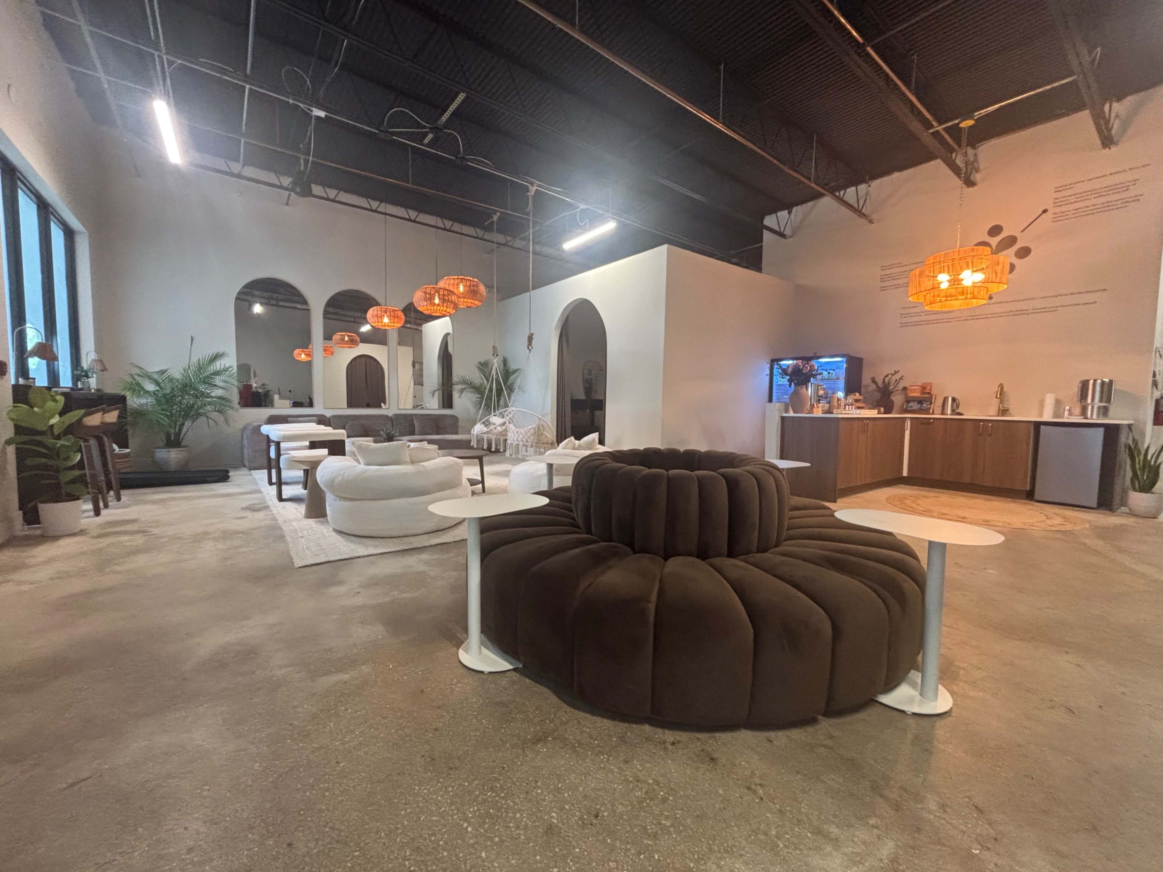 The image shows a modern lounge area with circular seating, potted plants, and pendant lights, alongside a kitchenette and large windows.
