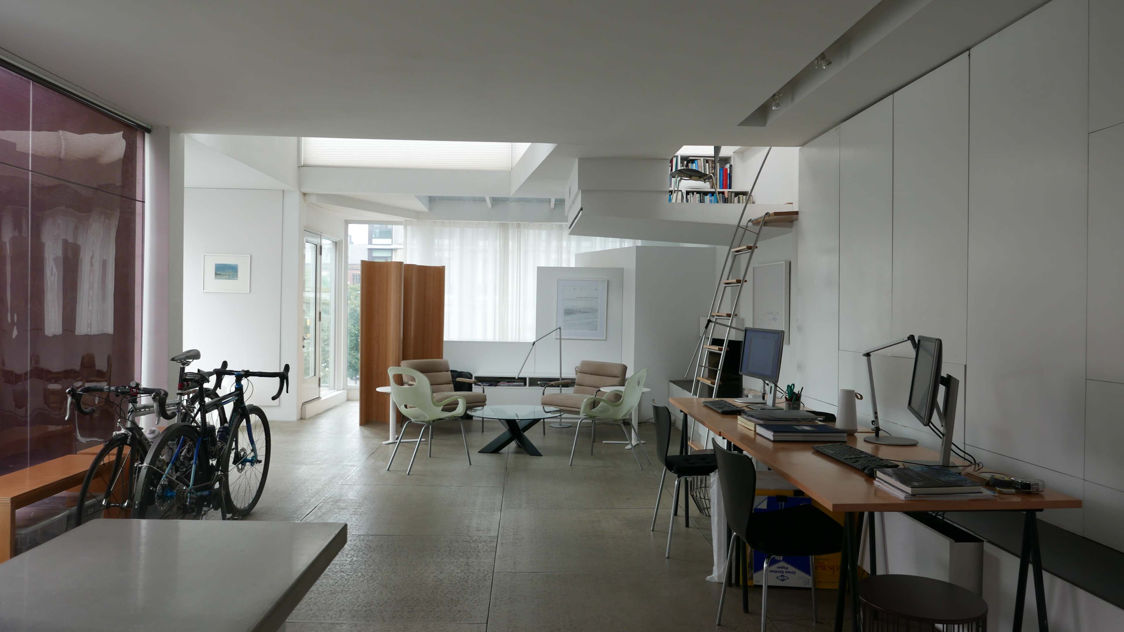 A modern, open-concept living space featuring a dining area, workspace with computers, and bicycles leaning against a wall.