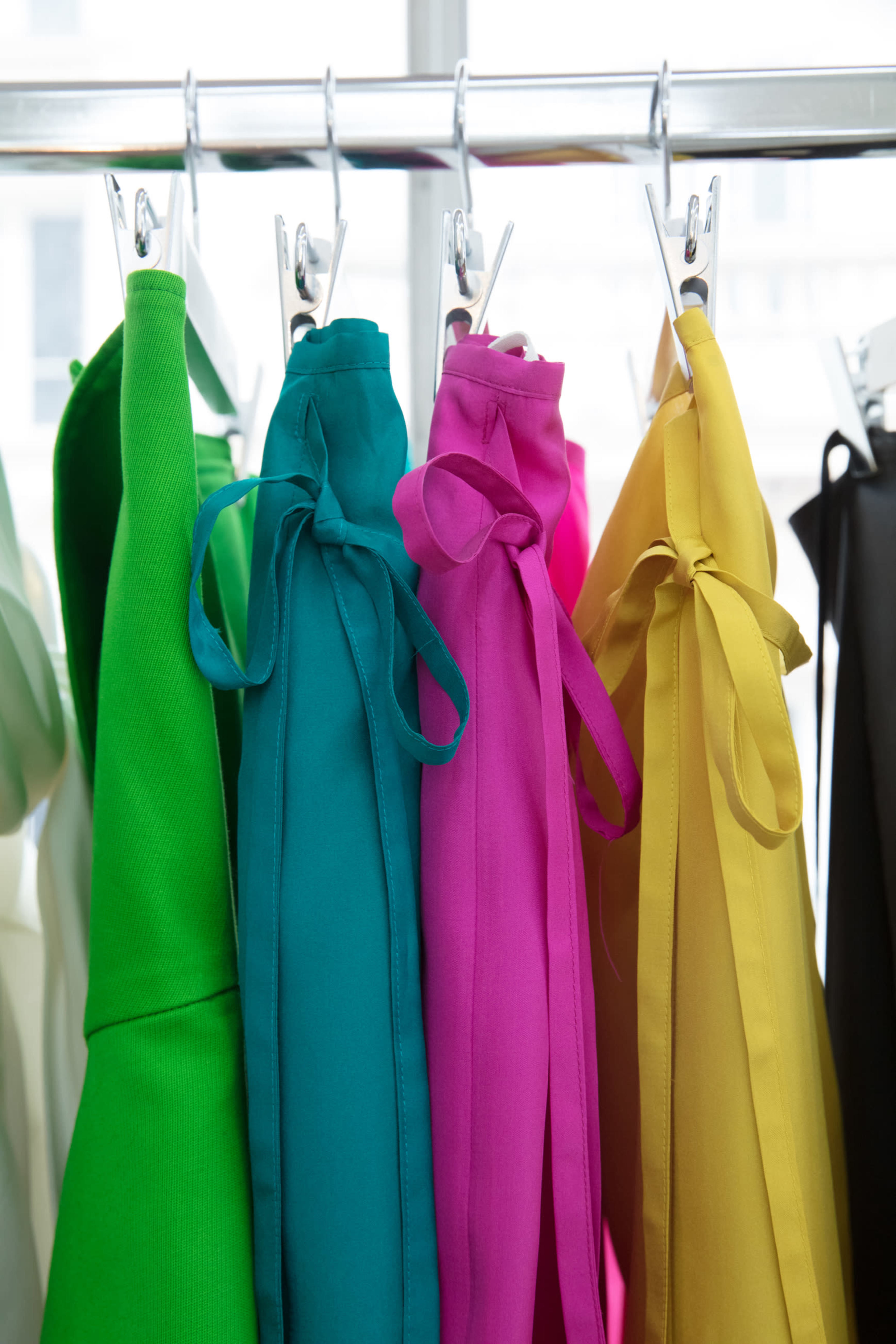 Several dresses in vibrant colors, including green, teal, pink, and yellow, hang on a rack with tied belts.