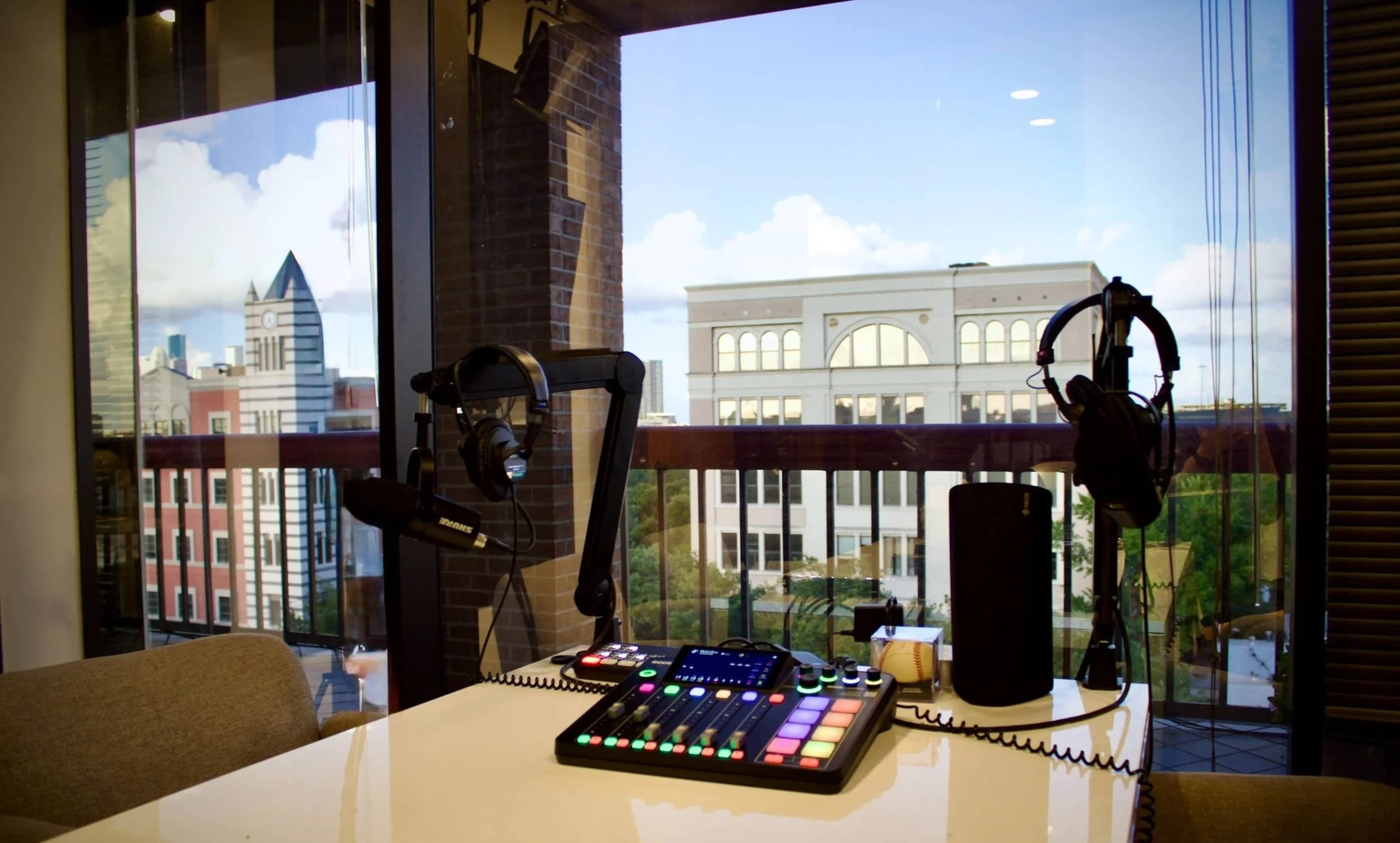 The image shows a modern recording setup with a soundboard, microphones, headphones, and a view of buildings through a large window.