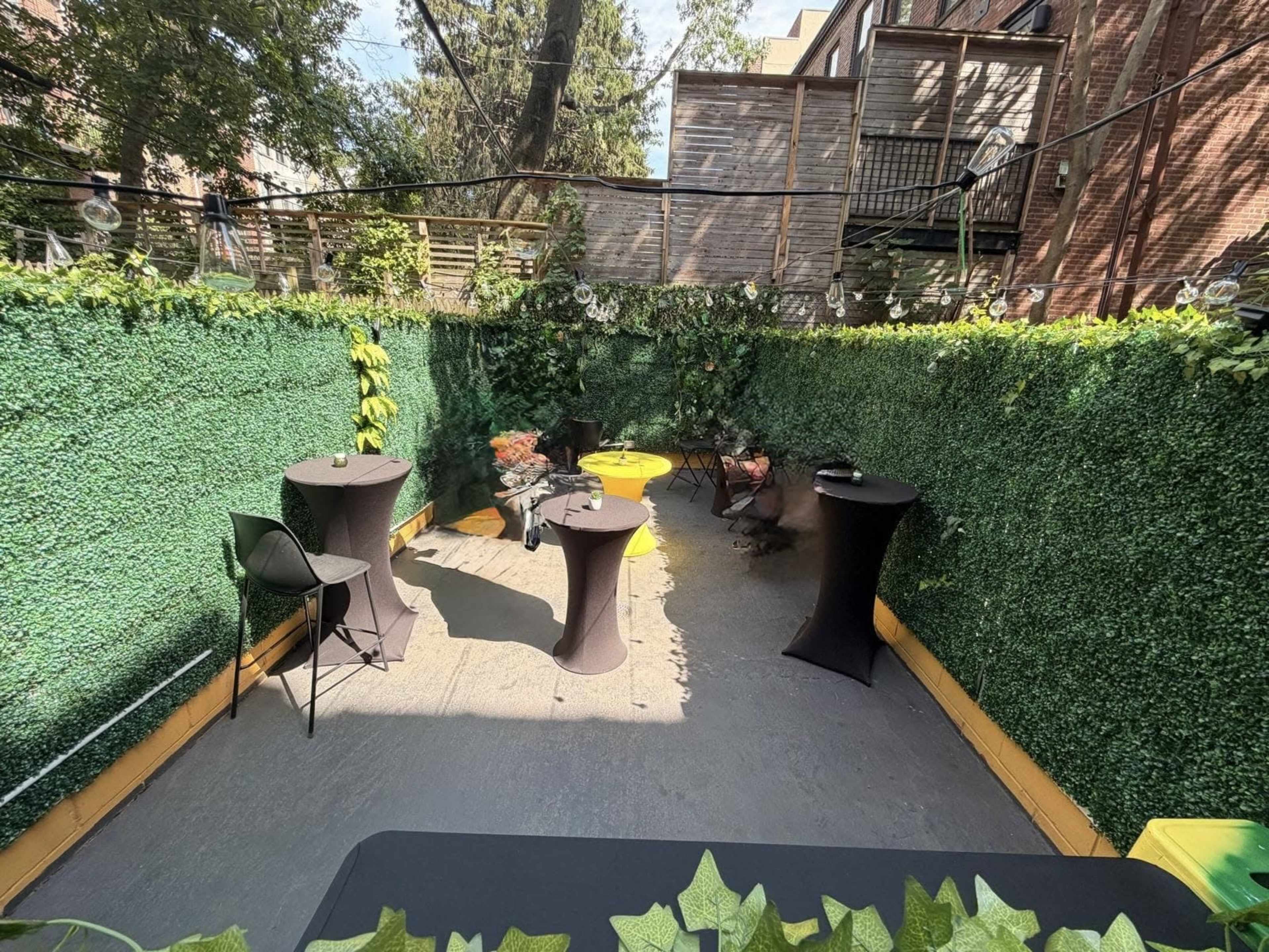 The image shows an outdoor space decorated with artificial greenery, featuring black cocktail tables and a few chairs arranged along the perimeter.