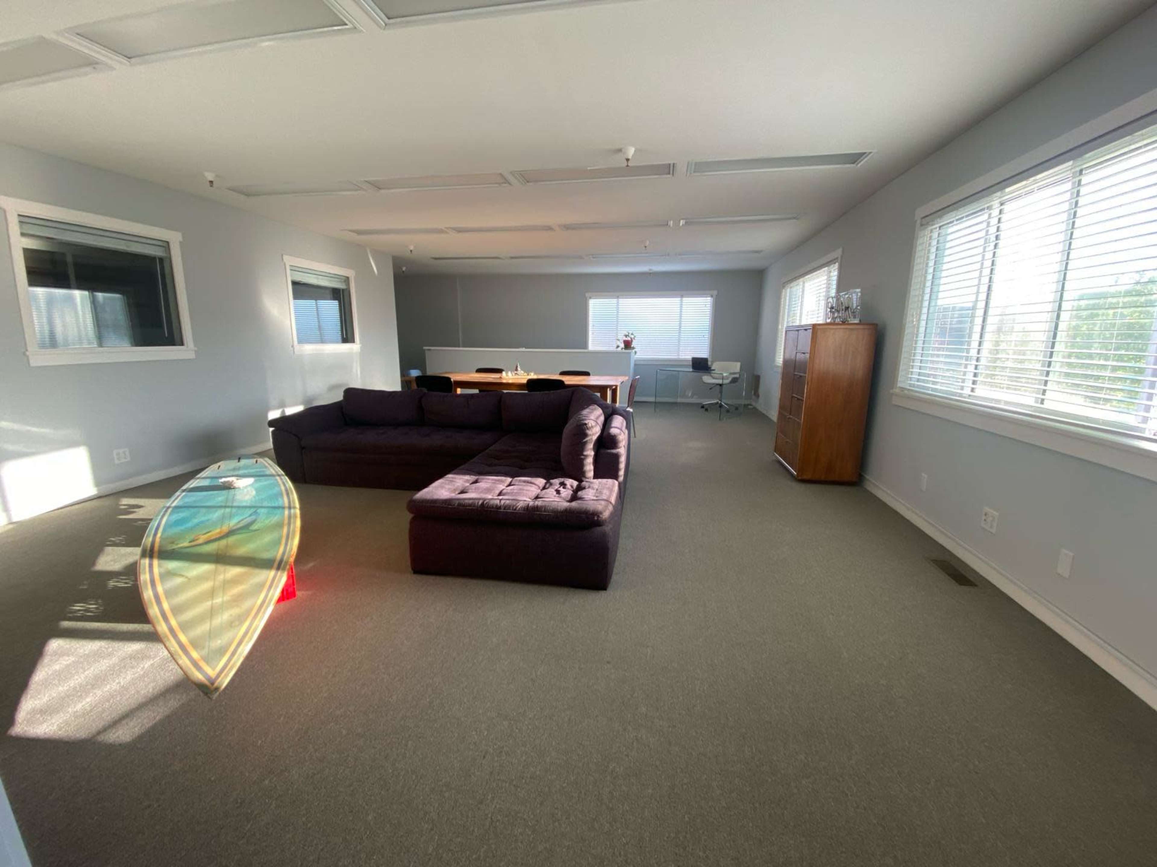 The image shows a spacious, modern office lounge area with a gray carpet, a dark sectional sofa, a glass coffee table, and large windows providing natural light.