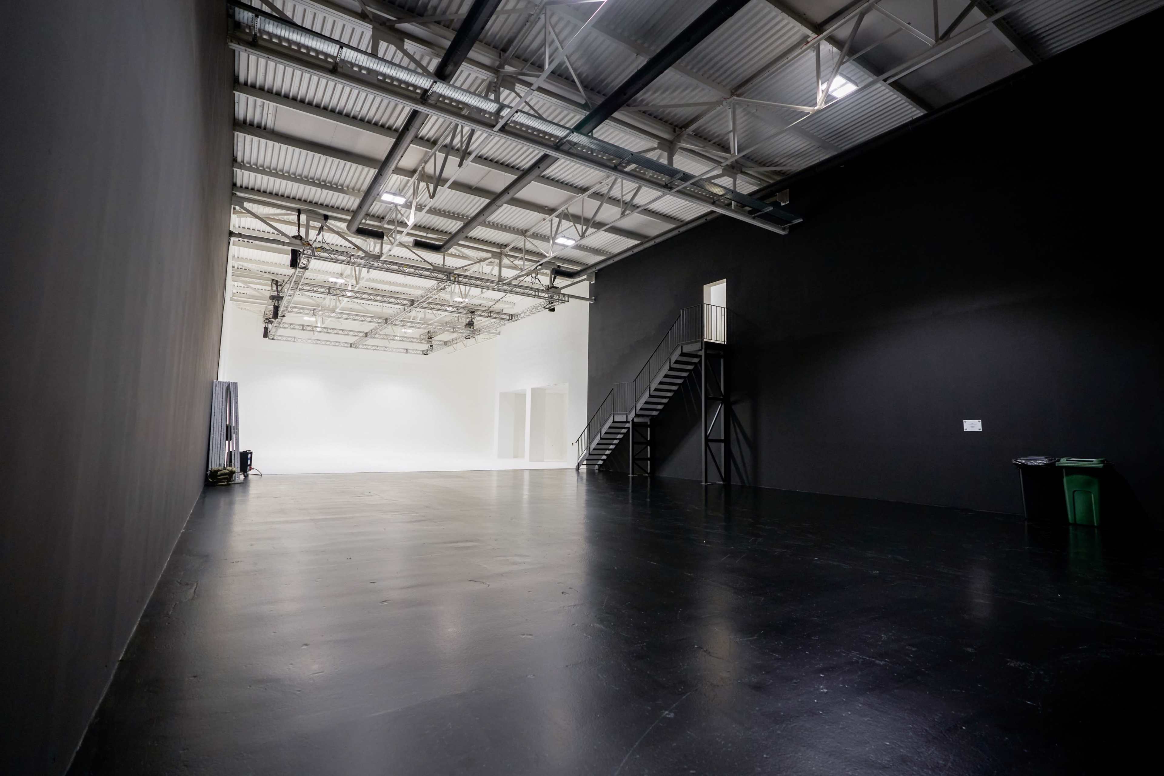 The image shows a large, empty industrial space with black walls, a polished floor, and a staircase leading to a second level.